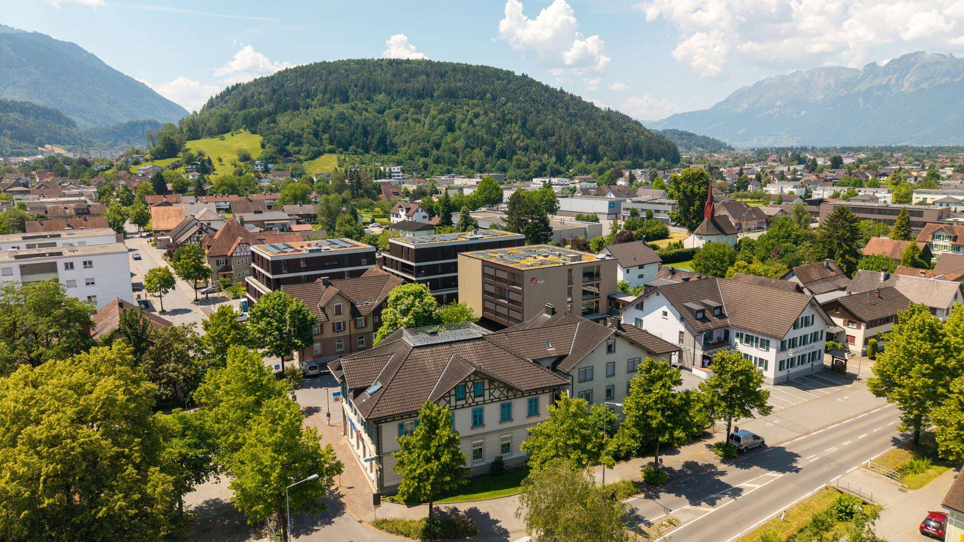 Panoramablick aus der Vogelperspektive auf eine Stadt mit bewaldeten Hügeln und Bergen.