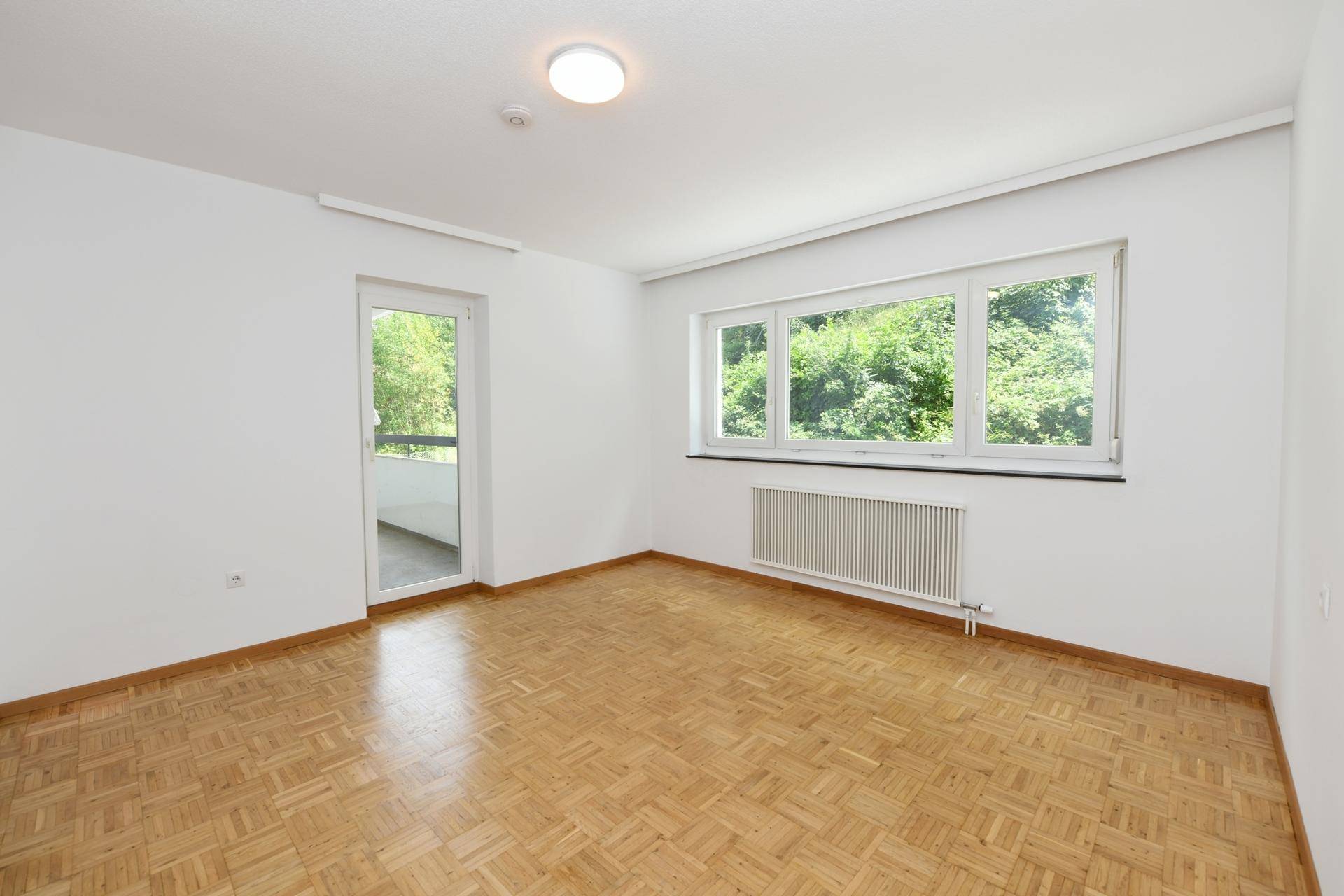Leerer Raum mit Fischgrätparkett, großem Fenster und Zugang zu einem Balkon.