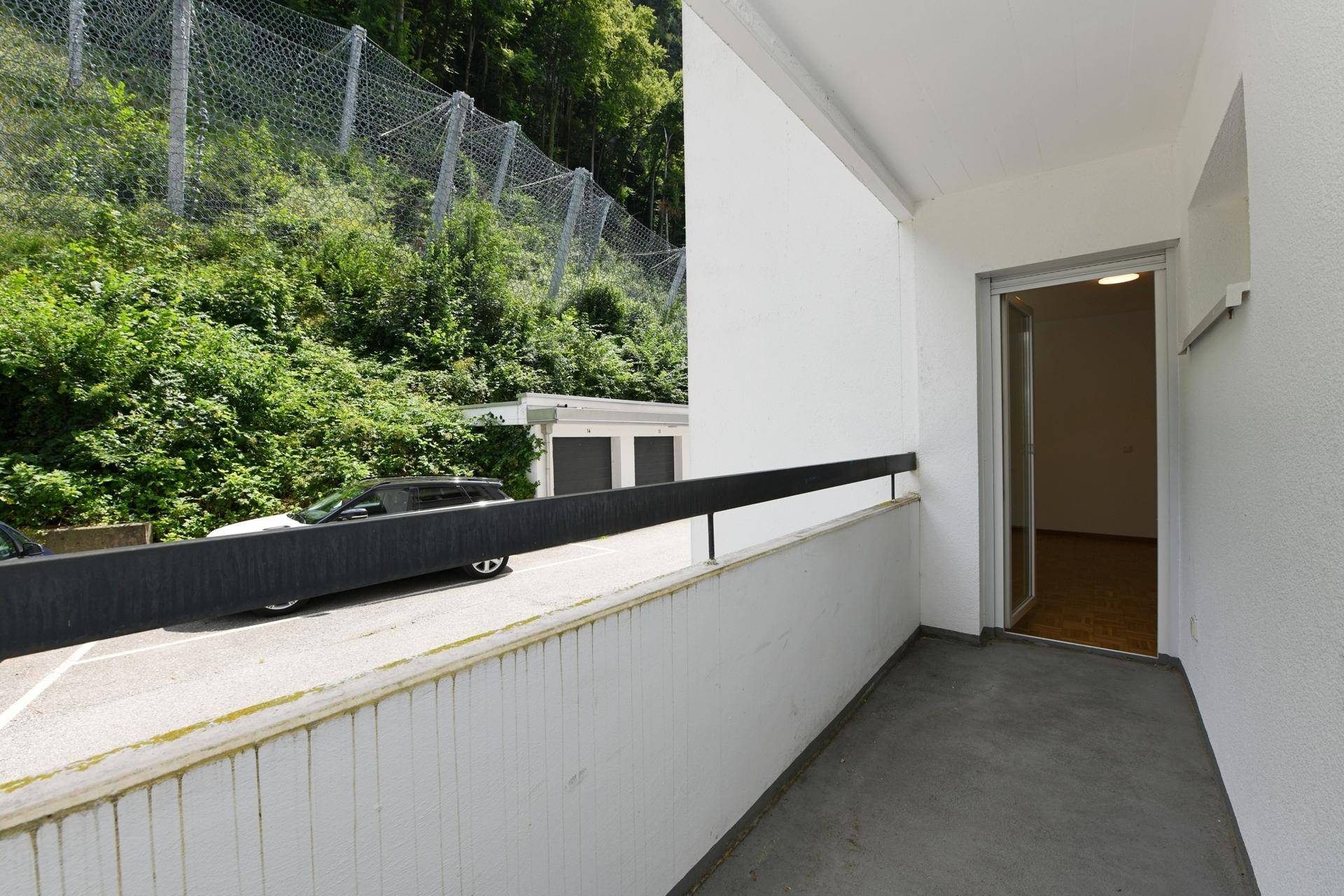 Balkon mit Zugang zur Wohnung und Aussicht auf eine bewaldete Anhöhe und Parkplätze.