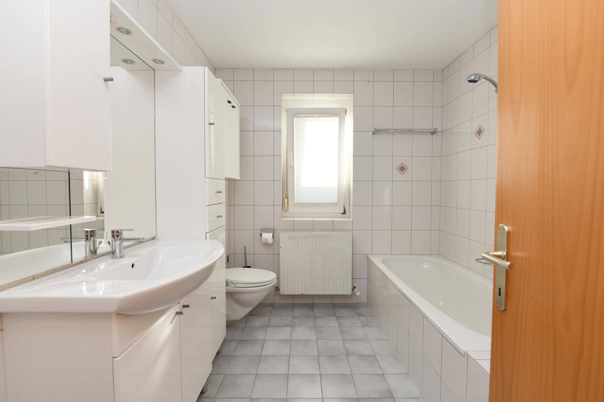 Helles Badezimmer mit weißen Fliesen, Badewanne, Waschbecken und Fenster.
