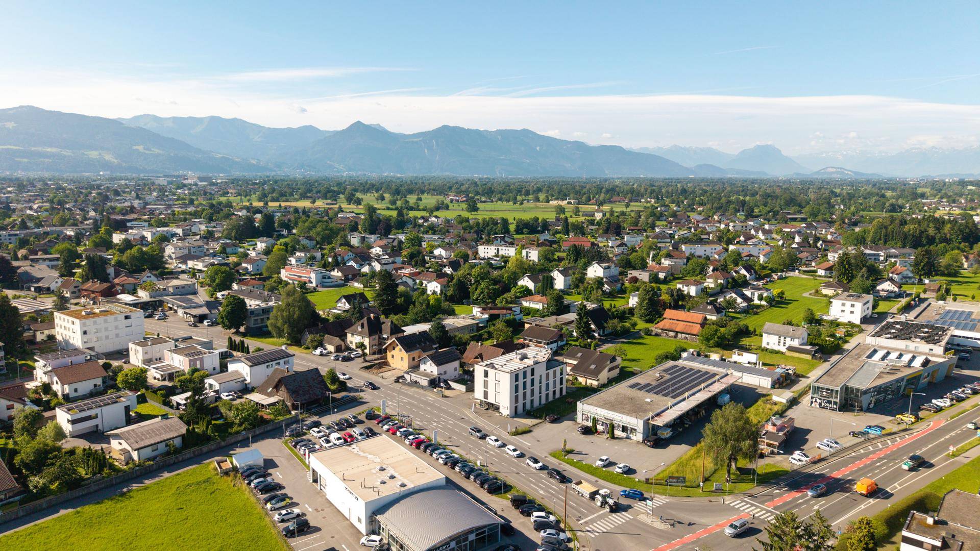 Weitläufige Luftaufnahme einer Stadt mit beeindruckender Bergkulisse im Hintergrund und grünen Flächen.