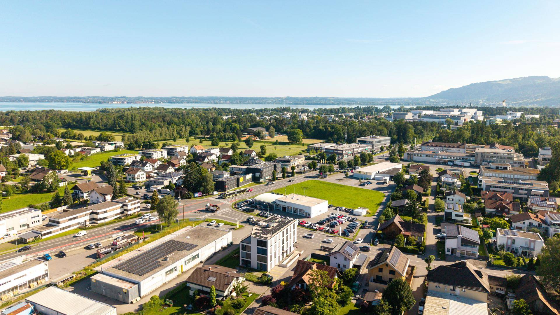 Panoramablick aus der Luft auf eine Stadt mit See und Bergen am Horizont unter blauem Himmel.