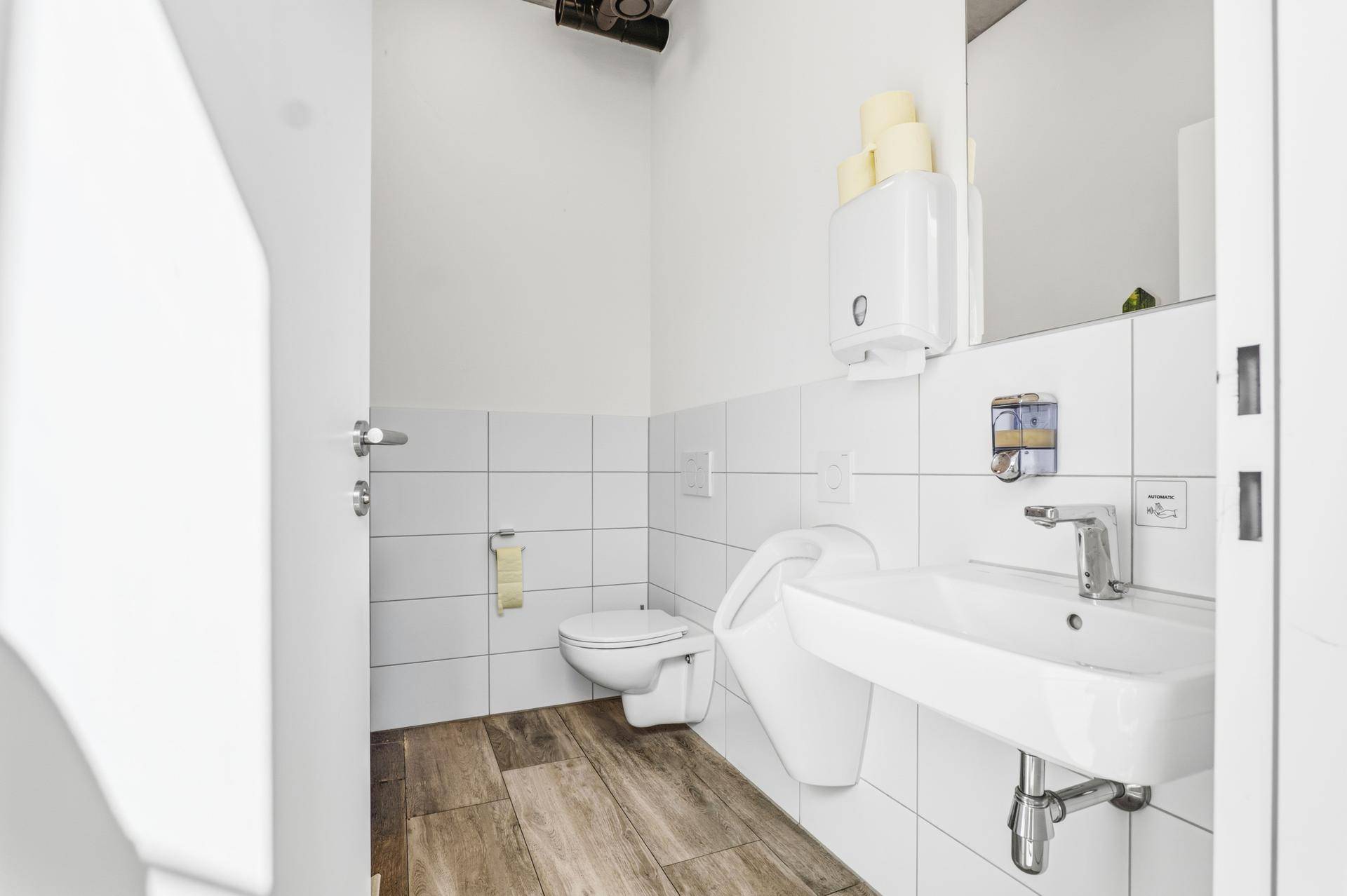 Moderne Toilette mit Urinal, Waschbecken und weißen Fliesen, die einen sauberen Eindruck macht.