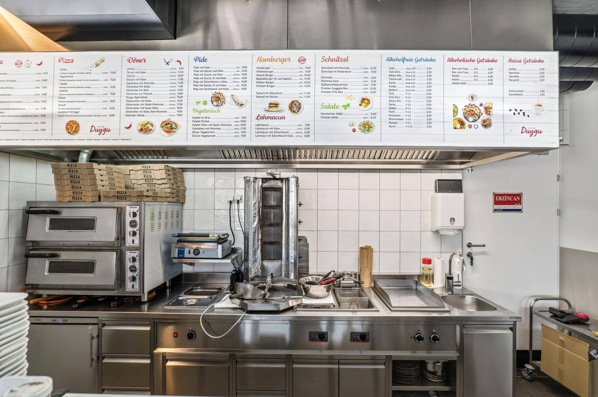 Professionelle Gastroküche mit Dönergrill, Pizzaöfen und Edelstahl-Arbeitsflächen unter der Speisekarte.