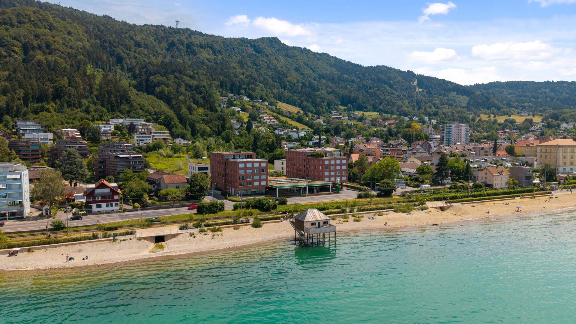 Panoramablick auf die Gebäude am Seeufer mit Strand und bewaldeten Hügeln.