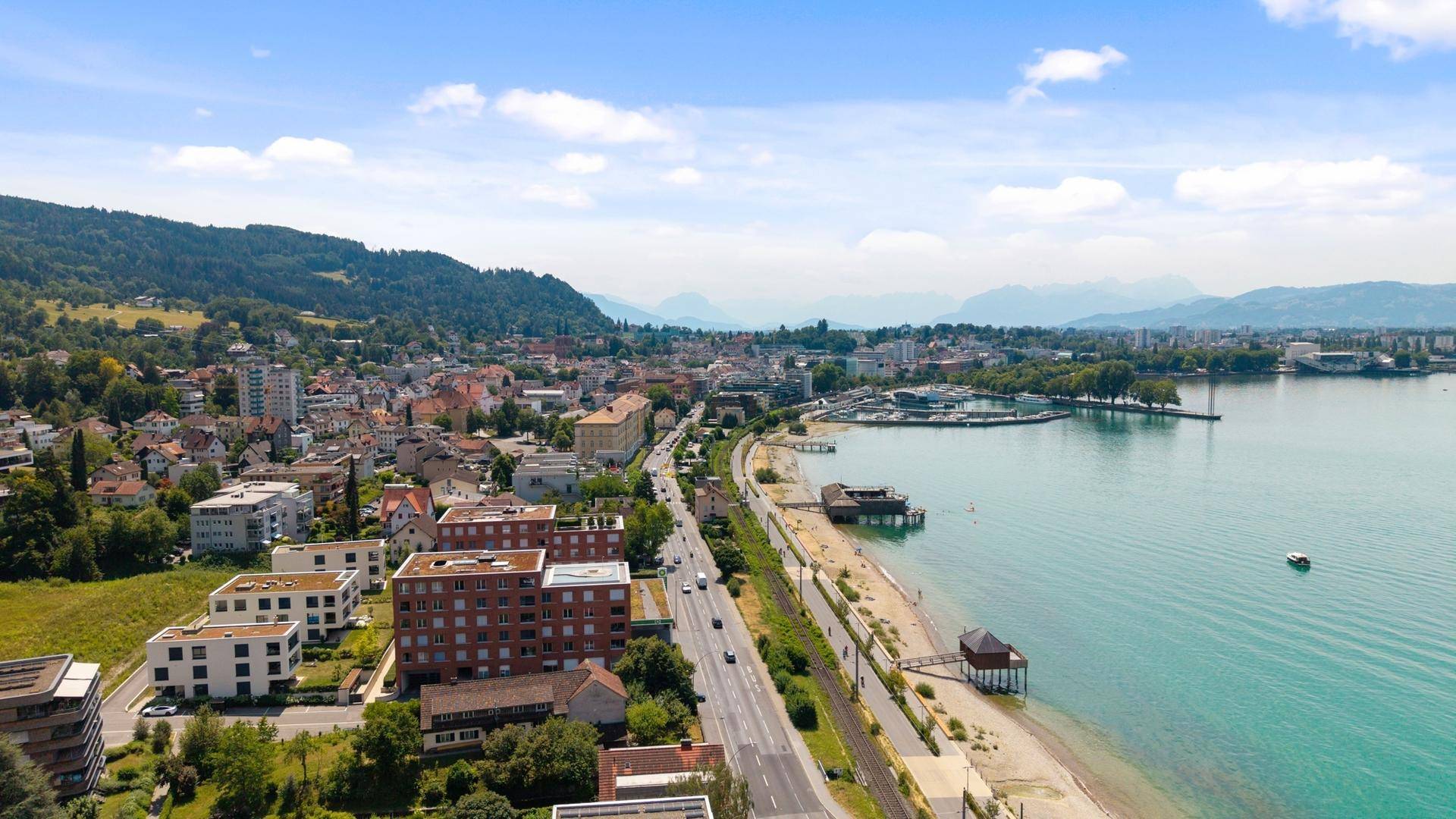 Stadtansicht entlang des Seeufers mit Gebäuden, Hafen und Bergen im Hintergrund.