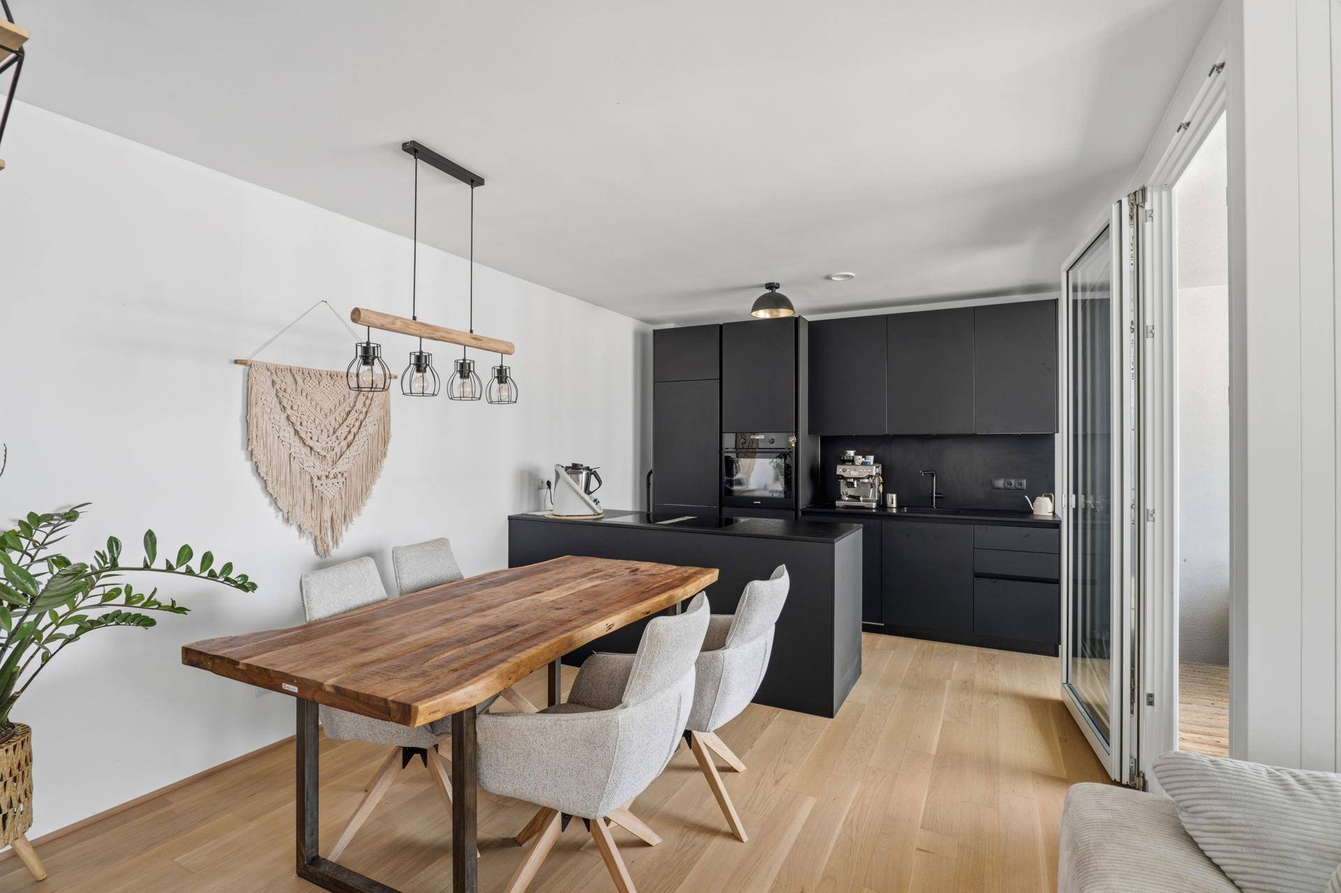 Offener Wohn- und Essbereich mit schwarzer Küche, Holztisch und Zugang zum Balkon.