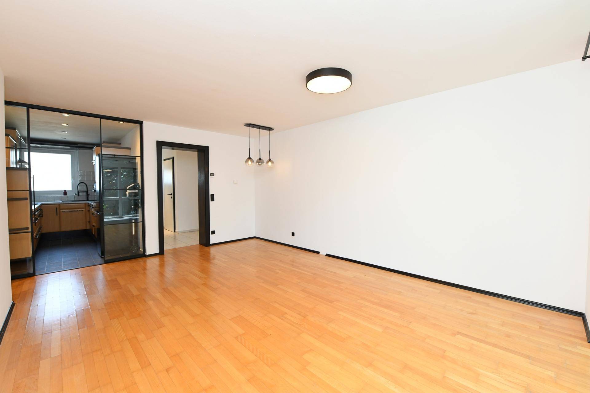 Geräumiger Wohnbereich mit Holzboden und Zugang zur Küche durch eine moderne Glaswand.