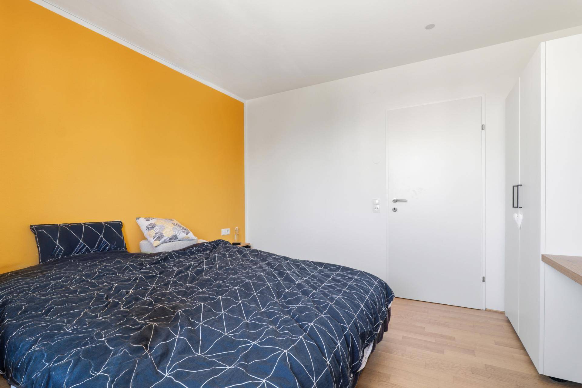 Schlafzimmer mit gelber Akzentwand, Holzboden und weißer Tür.