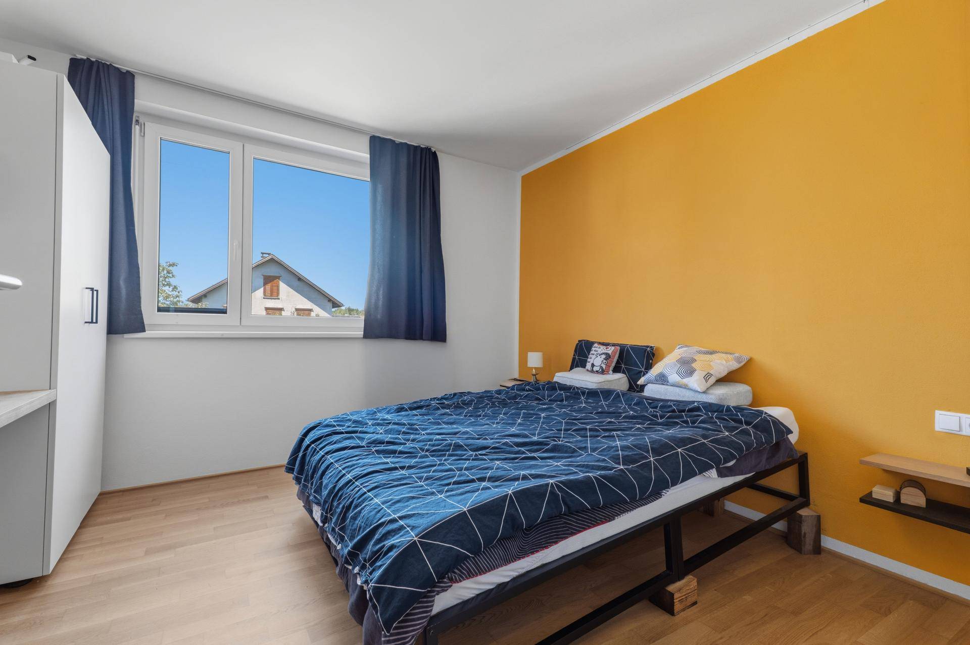 Schlafzimmer mit gelber Akzentwand, Holzboden und großem Fenster.