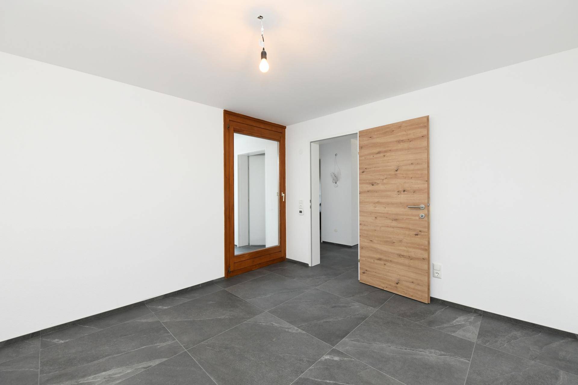 Unmöblierter Raum mit dunklen Fliesen und Holztür, bereit zur individuellen Einrichtung.