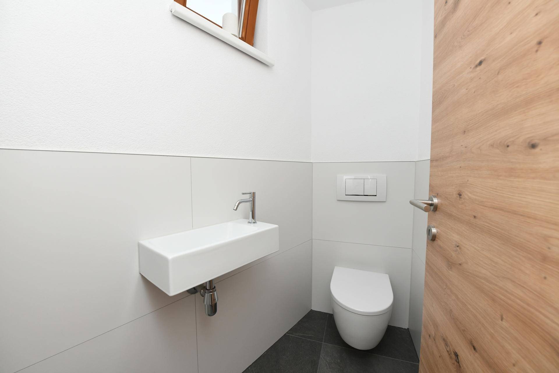 Gäste-WC mit wandhängendem WC, kleinem Waschbecken und einem Fenster für Belüftung.