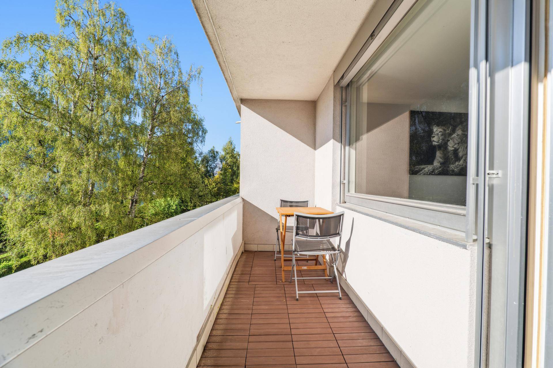 Schmaler Balkon mit Holzboden, Sitzgelegenheiten und Blick auf Bäume.