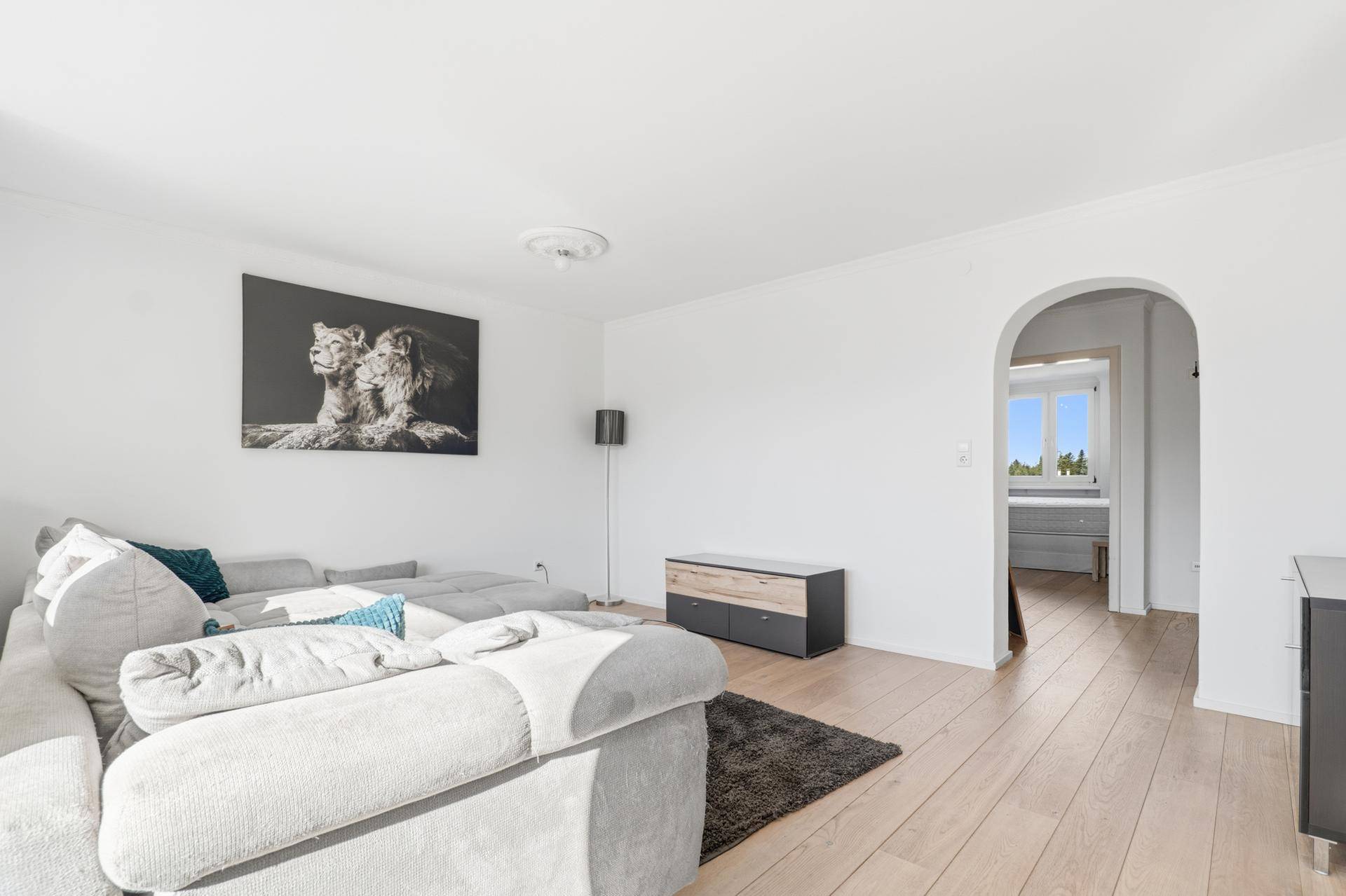 Geräumiges Wohnzimmer mit hellem Holzboden und einem Durchgang zu einem weiteren Raum.