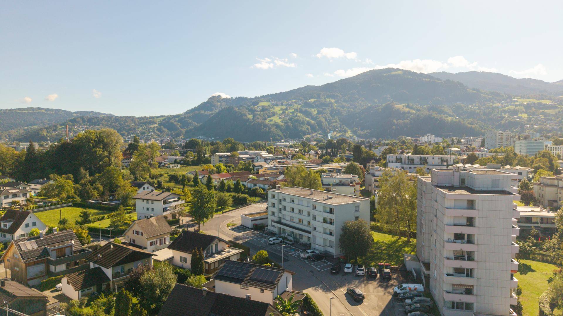 Luftaufnahme einer Wohnsiedlung mit grünen Hügeln und Bergen im Hintergrund.