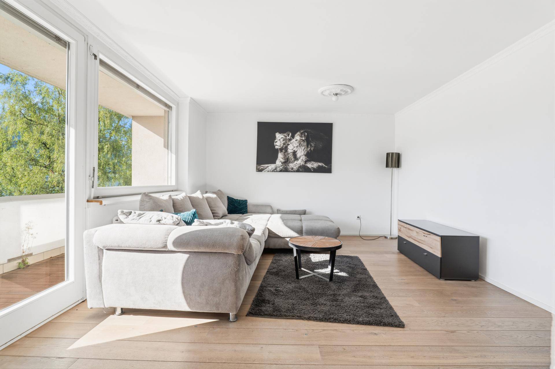 Helles Wohnzimmer mit Ecksofa, Holzboden und Zugang zu einem Balkon mit Grünblick.