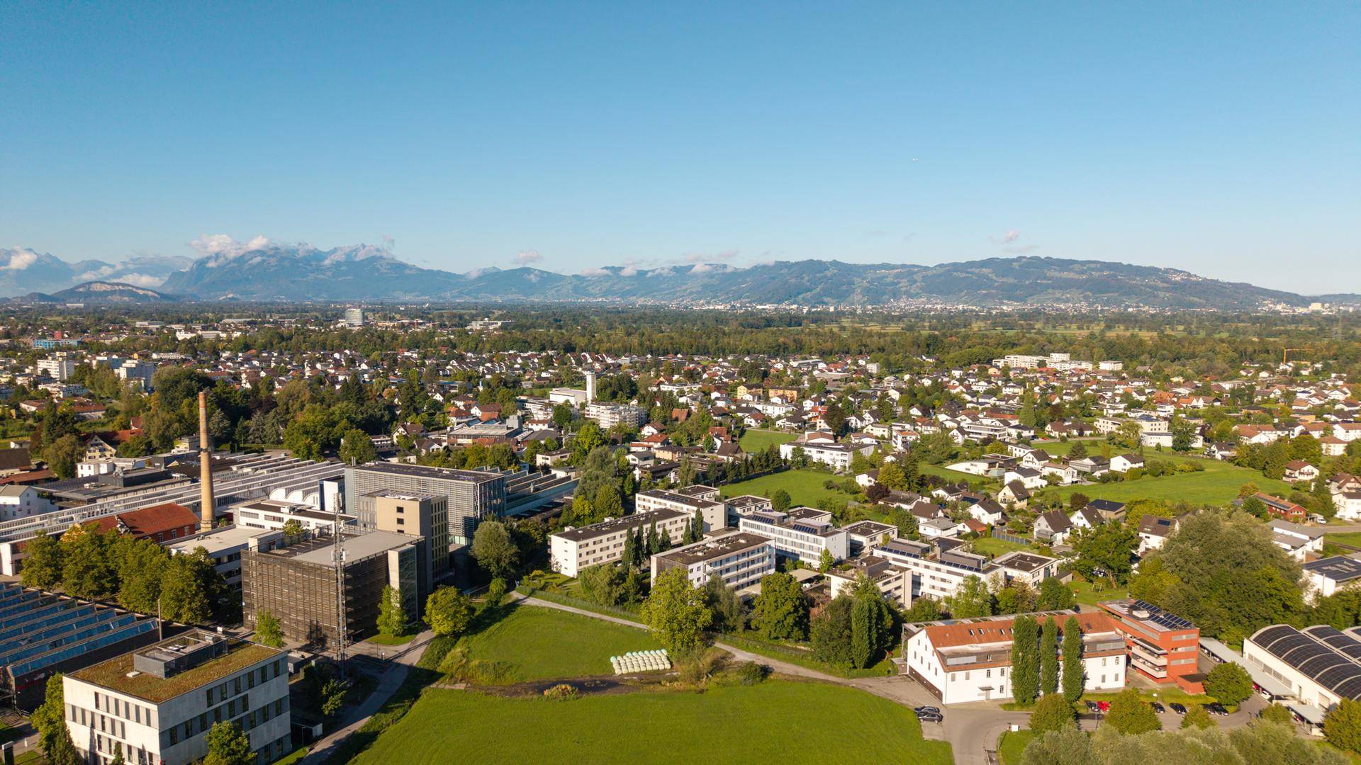 Weitläufige Luftaufnahme der Stadt mit grünen Flächen und Bergen im Hintergrund.