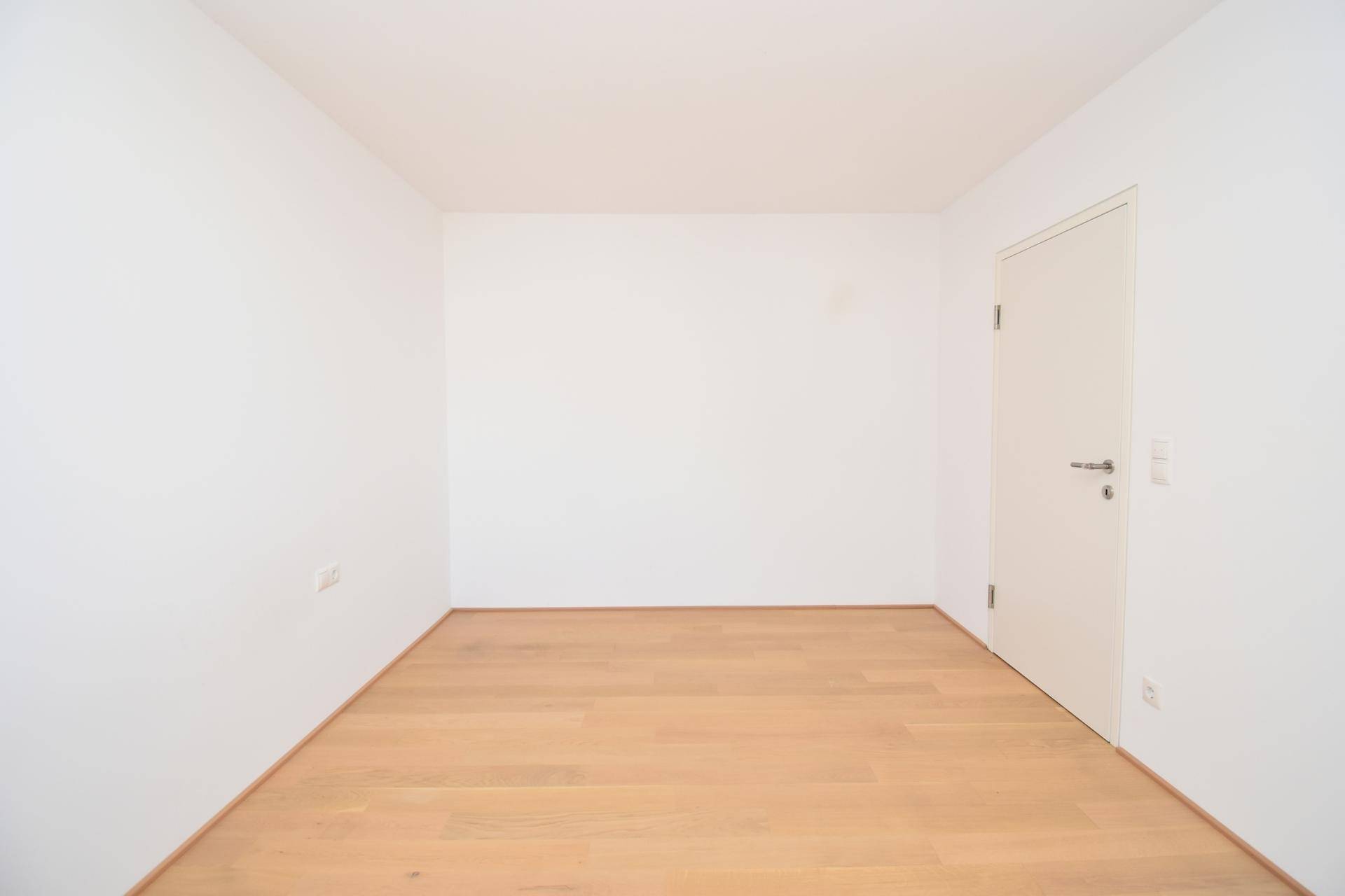 Leeres Zimmer mit hellem Parkettboden und weißen Wänden, ideal als Schlaf- oder Arbeitszimmer.