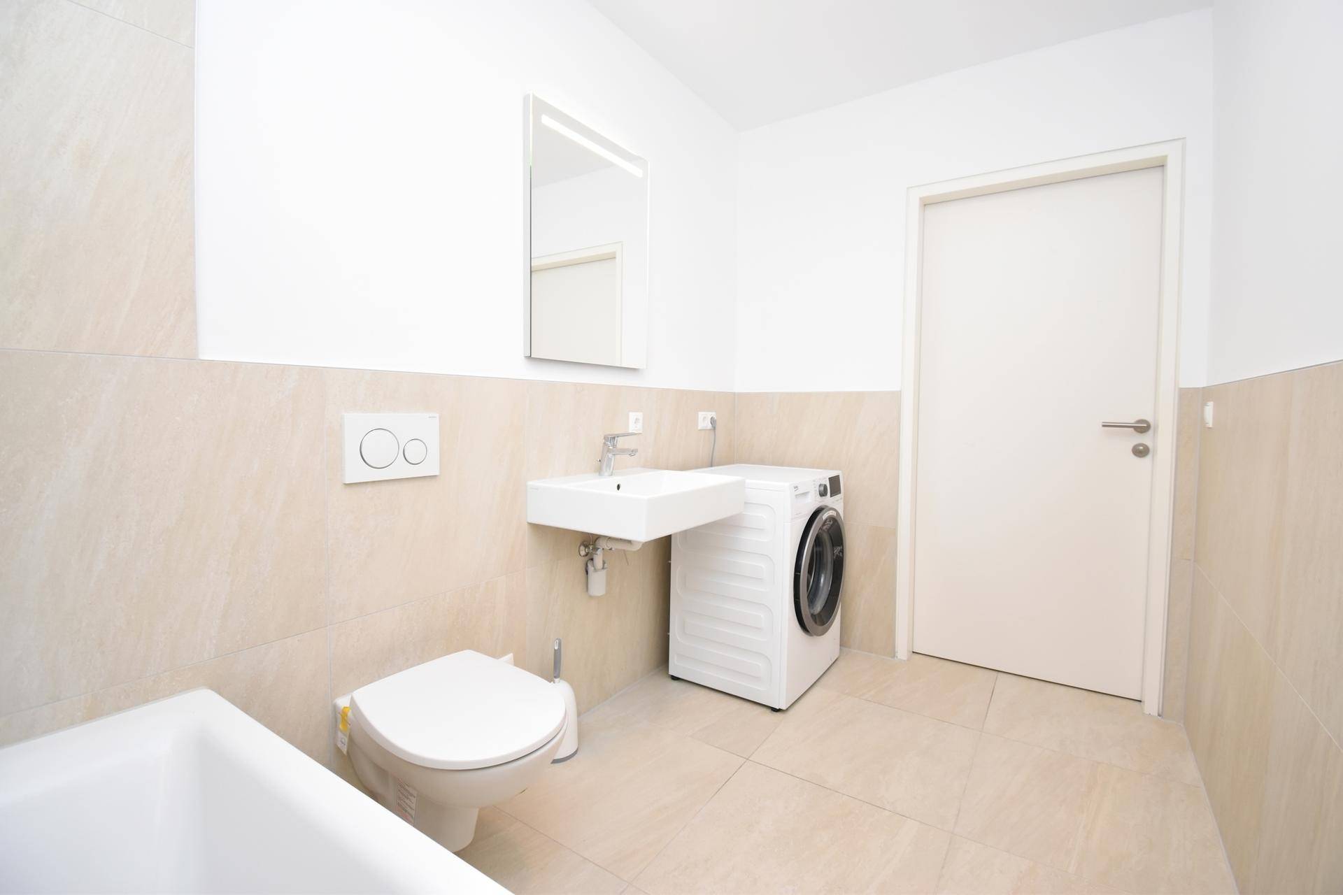 Helles Badezimmer mit Badewanne, wandhängendem WC und Waschmaschine, beige gefliest.