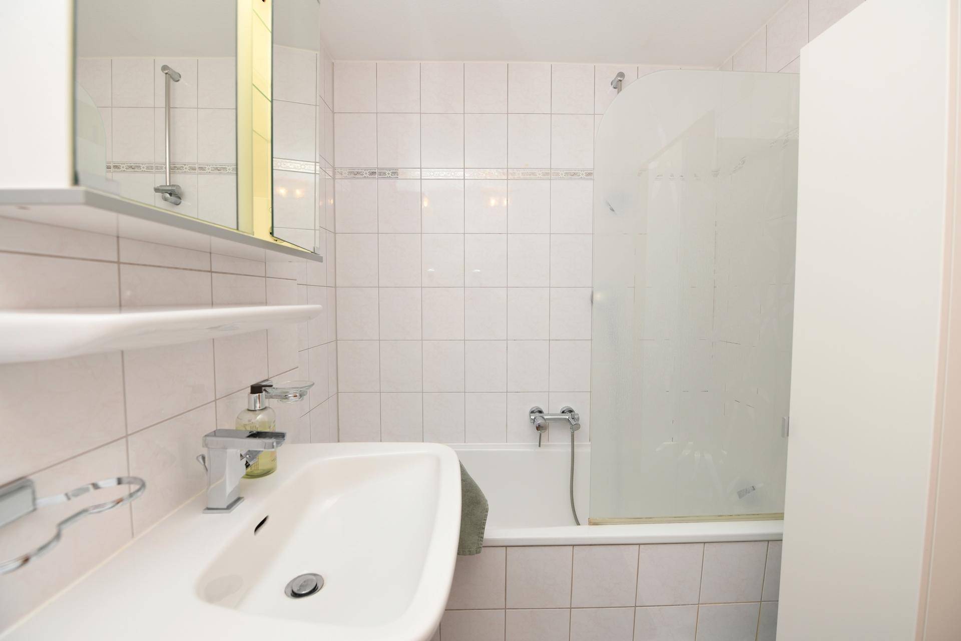 Helles Badezimmer mit weißen Fliesen, Waschbecken und Badewanne mit Duschwand.
