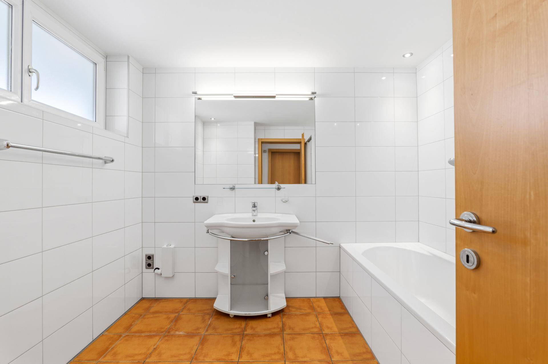 Badezimmer mit Badewanne, Waschbecken, Fenster und Terrakotta-Fliesenboden.