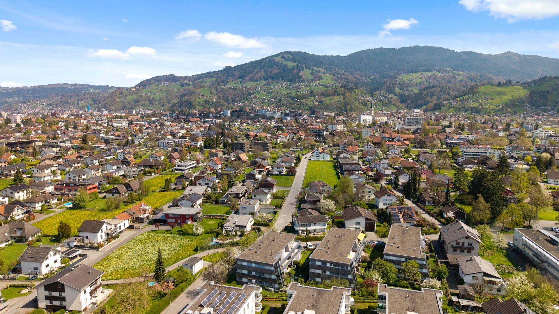 Panoramische Luftaufnahme einer Stadt mit Wohngebieten und bewaldeten Bergen.