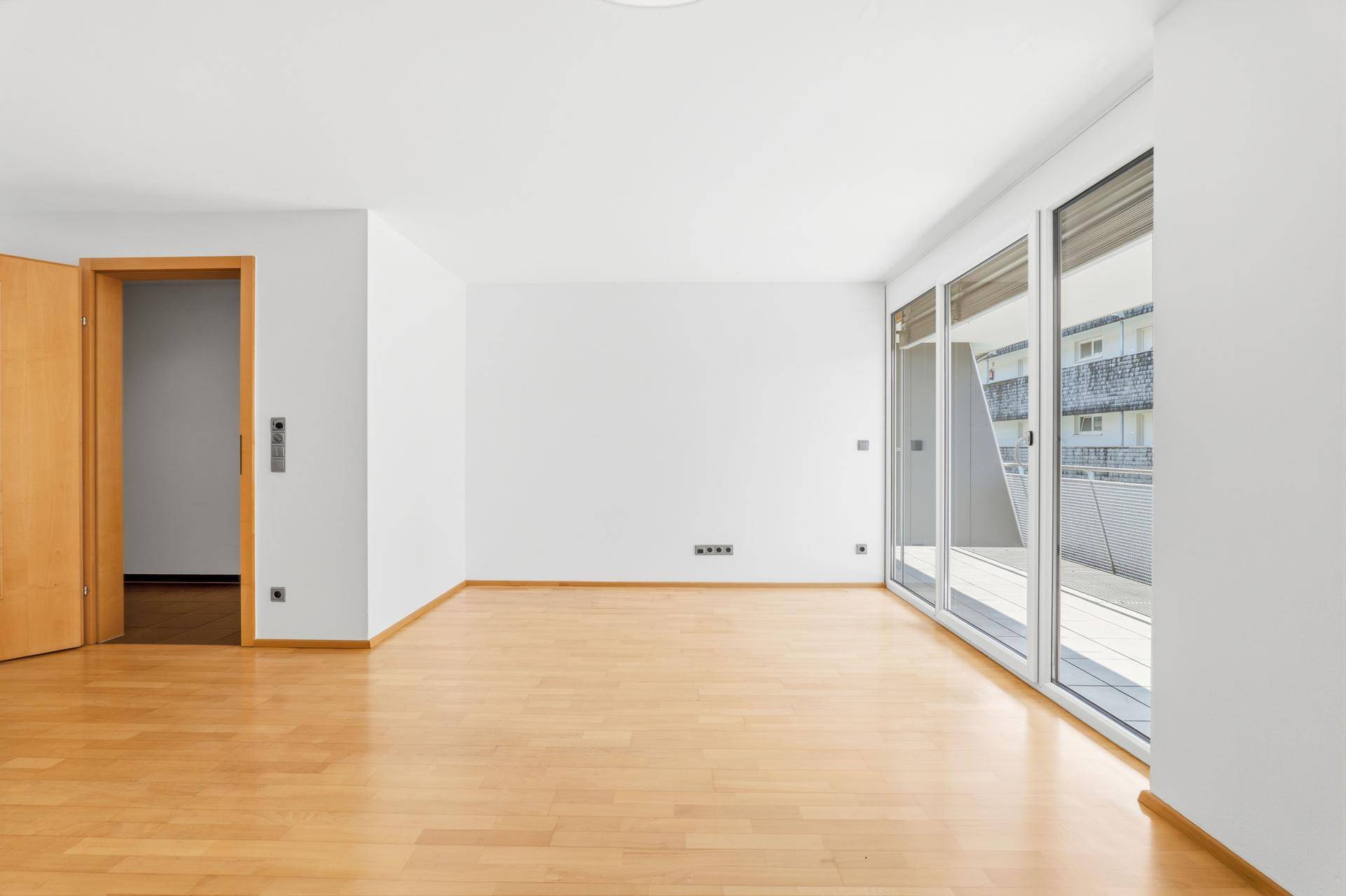 Geräumiges, leeres Wohnzimmer mit hellem Parkettboden und breiter Fensterfront.
