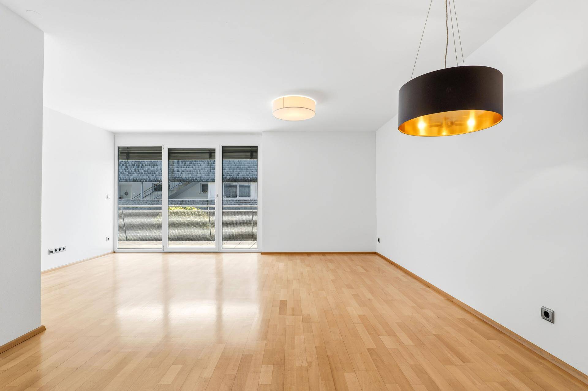 Helles, leeres Wohnzimmer mit Parkettboden und großen Fenstern, die auf einen Balkon führen.
