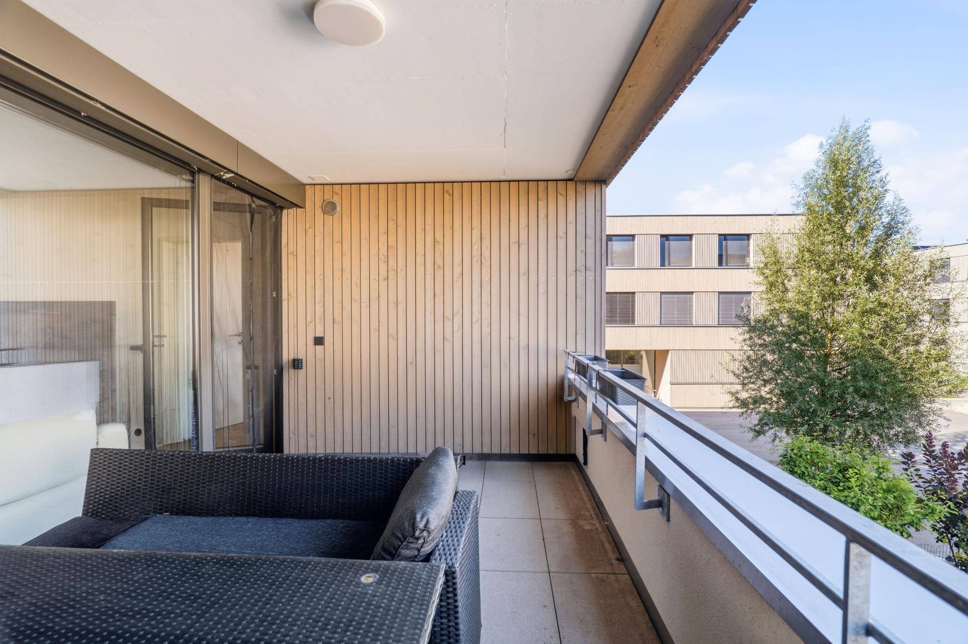 Balkon mit Holzwand, Sitzmöbeln und Blick auf die grüne Umgebung.