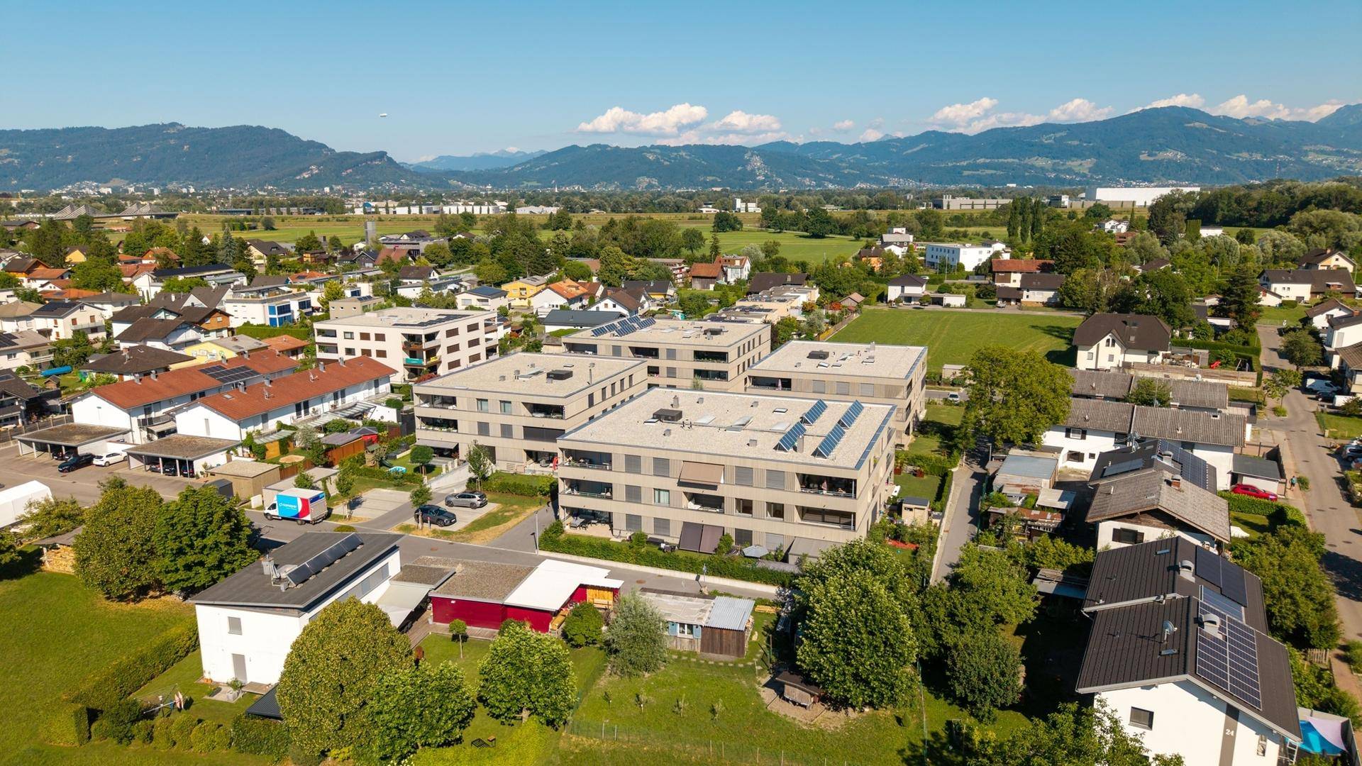 Luftaufnahme eines Wohngebiets mit modernen Gebäuden, grünen Flächen und Bergen im Hintergrund.