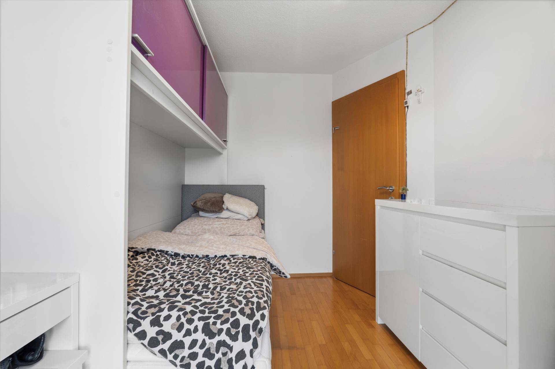 Kinderzimmer mit Einzelbett, lila Hängeschränken und weißer Kommode.