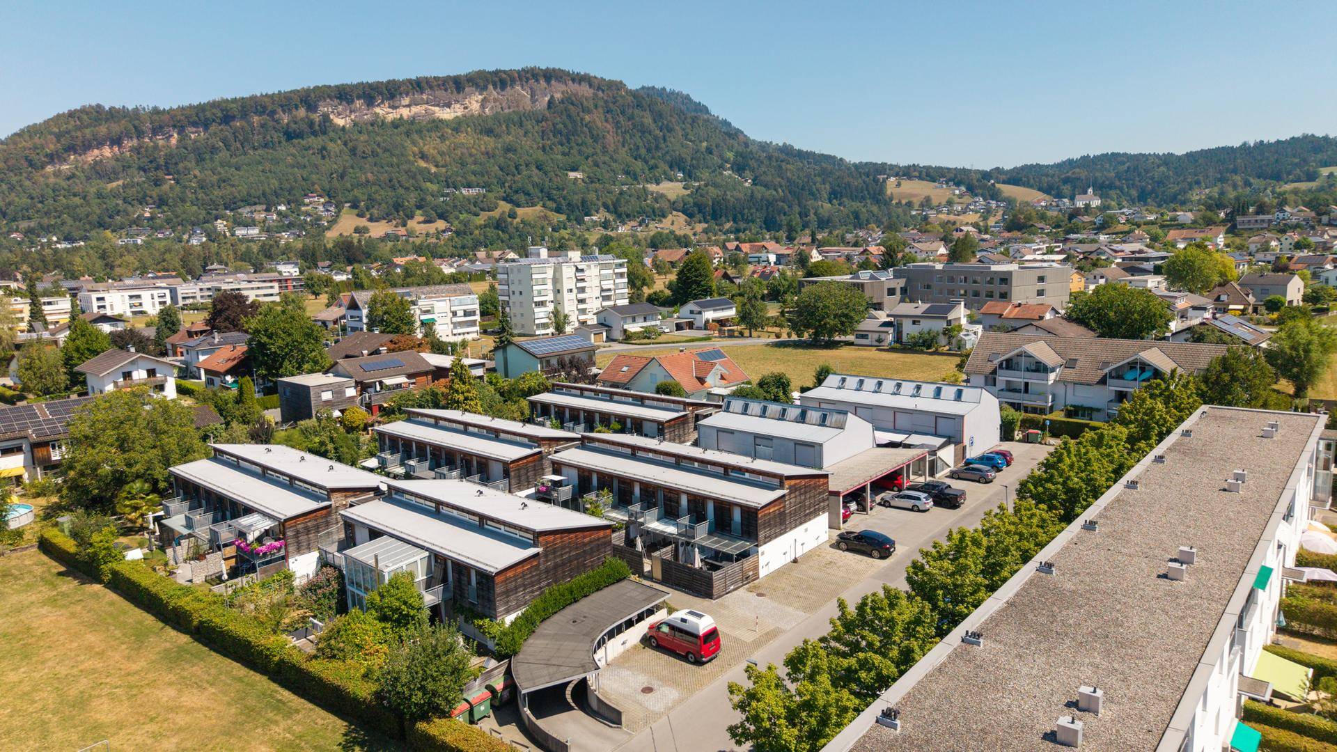 Luftaufnahme einer modernen Wohnanlage mit Holzelementen und einem bewaldeten Berg im Hintergrund.