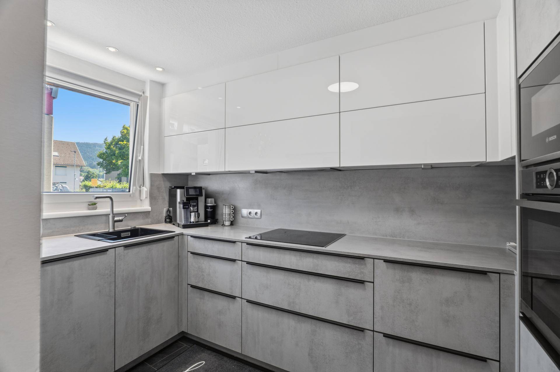 Moderne Küche mit grauen Arbeitsplatten, weißen Hochglanzschränken und Fensterblick.