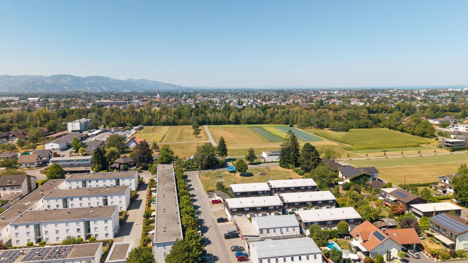 Panoramablick aus der Vogelperspektive auf eine Wohngegend mit Feldern und bewaldeten Hügeln am Horizont.