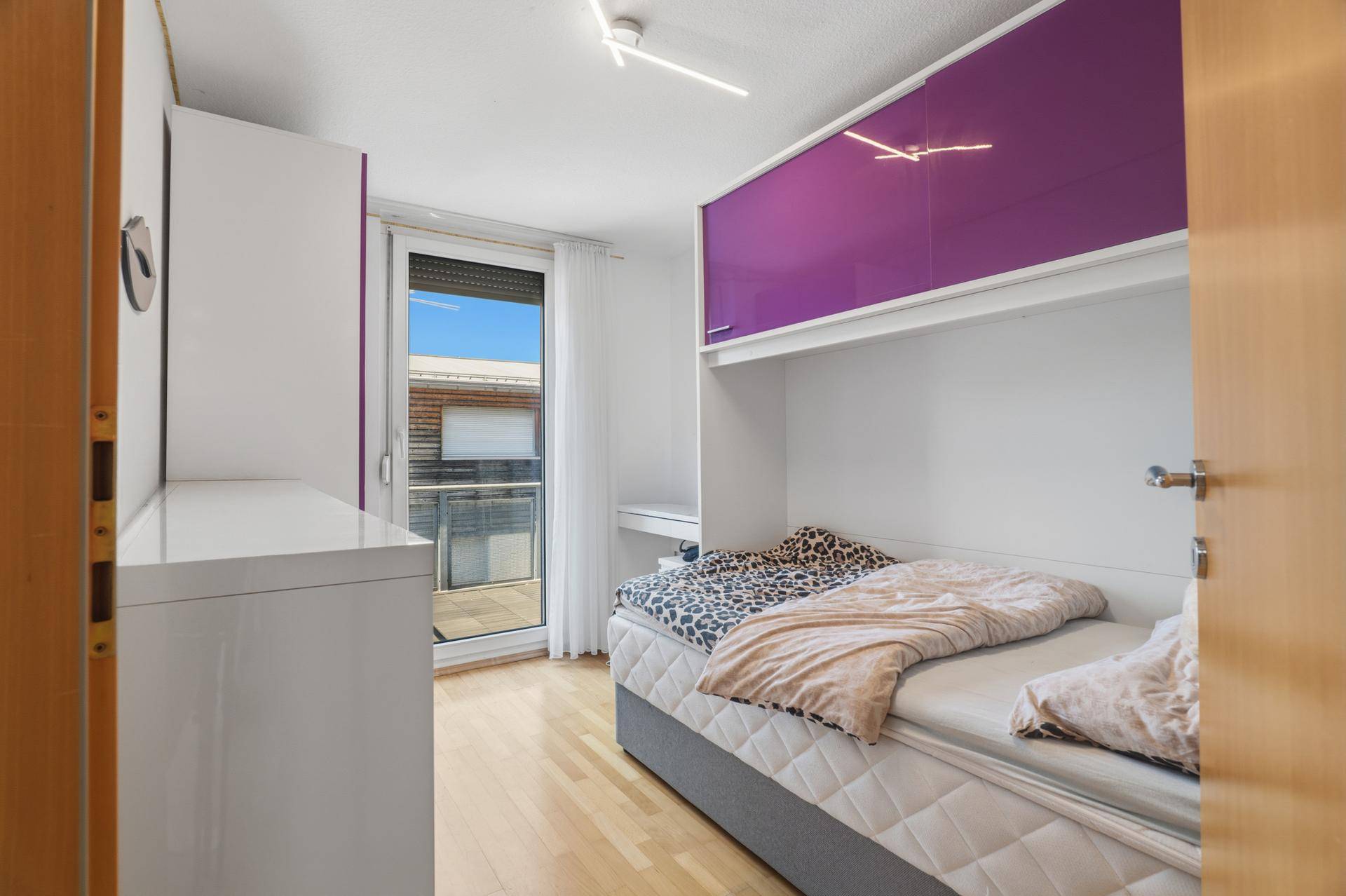 Kinderzimmer mit Einzelbett, lila Hängeschränken und Zugang zu einem Balkon.