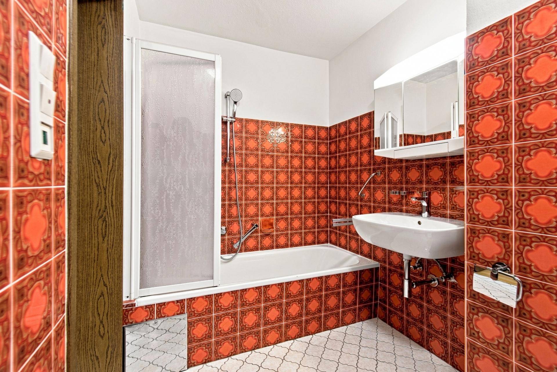 Retro-Badezimmer mit orangefarbenen Fliesen, Badewanne mit Duschwand, Waschbecken und WC, gut erhalten.