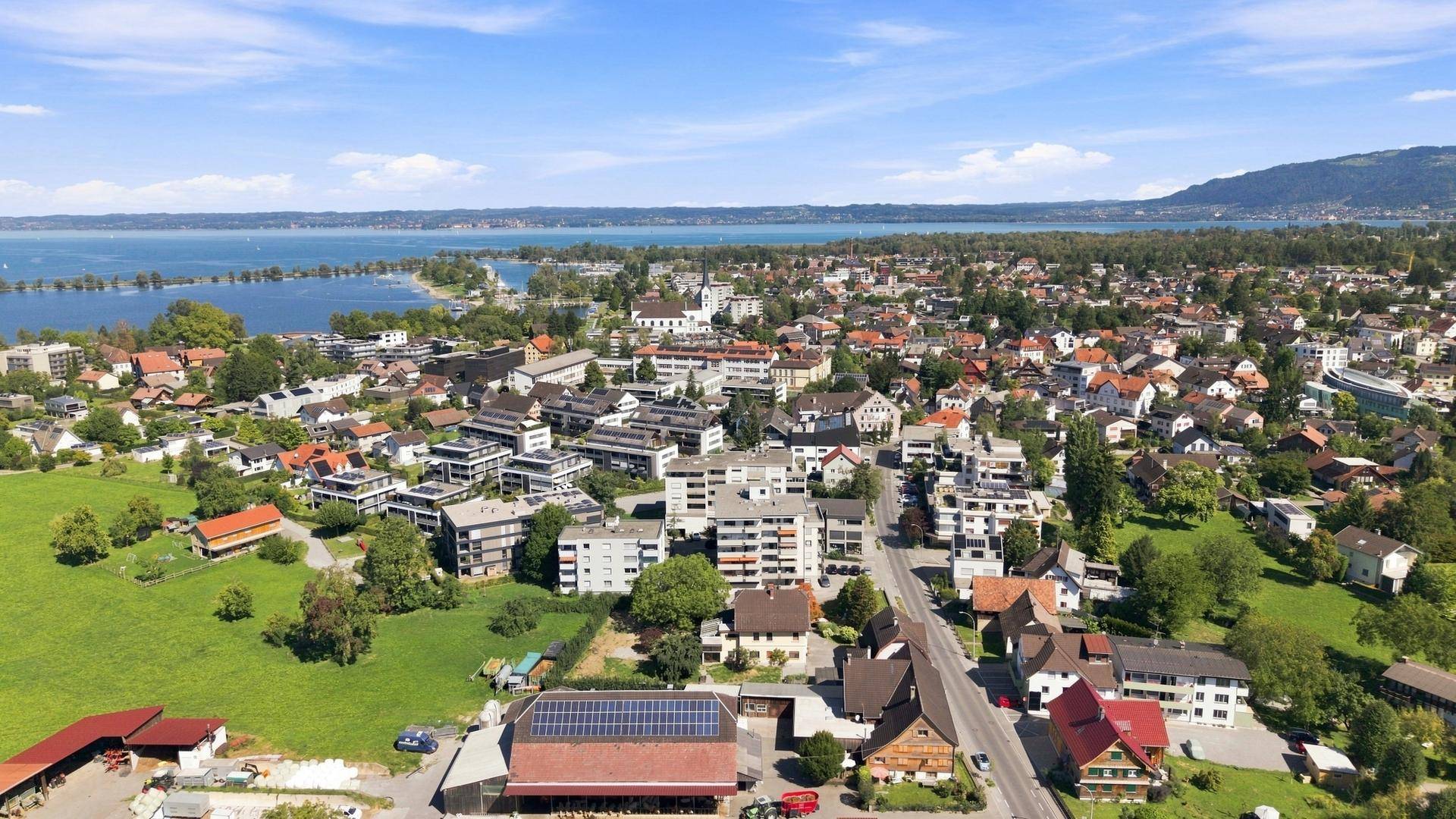 Luftaufnahme einer Wohnsiedlung am Seeufer mit grünen Feldern und klarem Himmel.