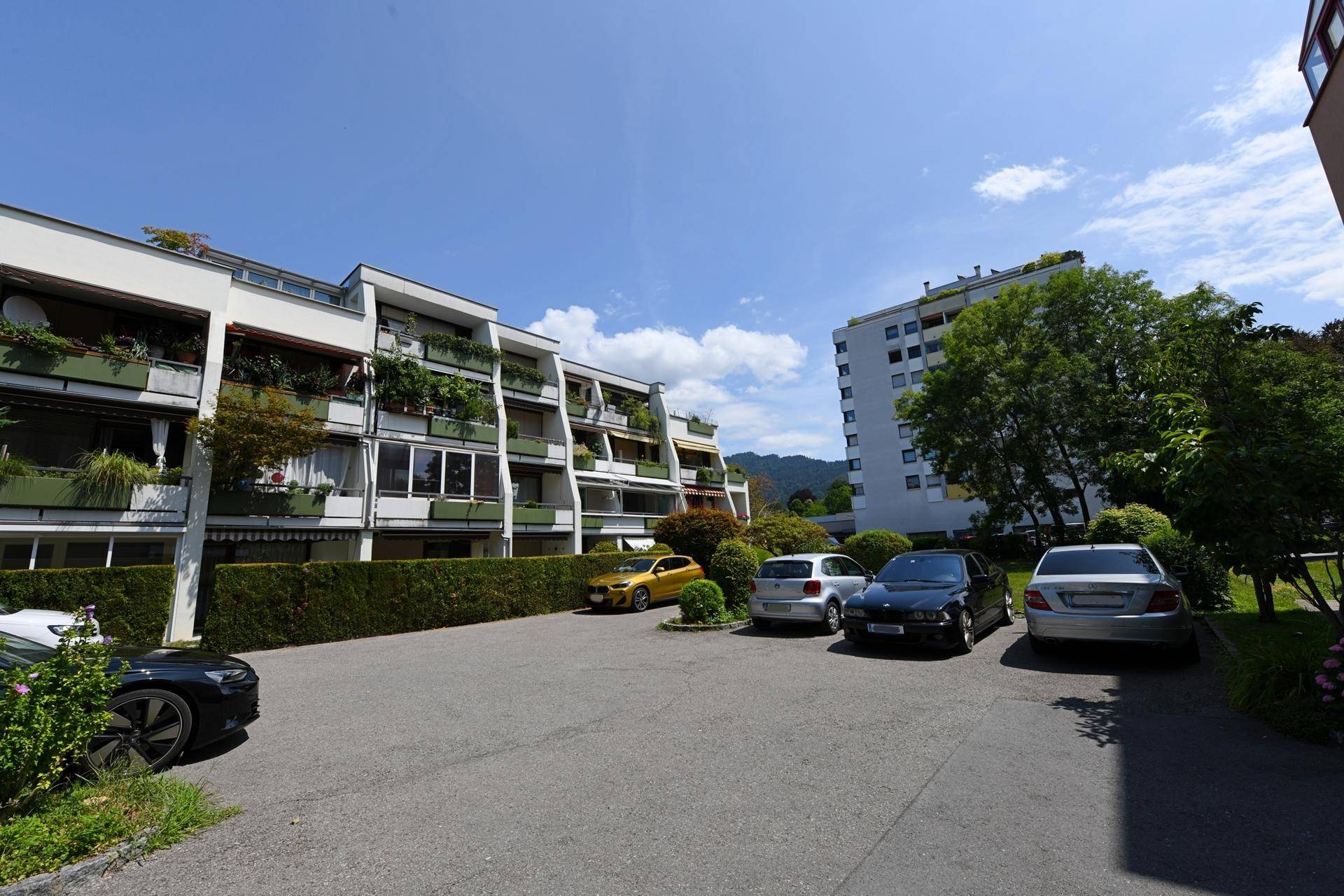 Außenansicht der Wohngebäude mit Parkplätzen, grüner Umgebung und Bergblick in der Ferne.