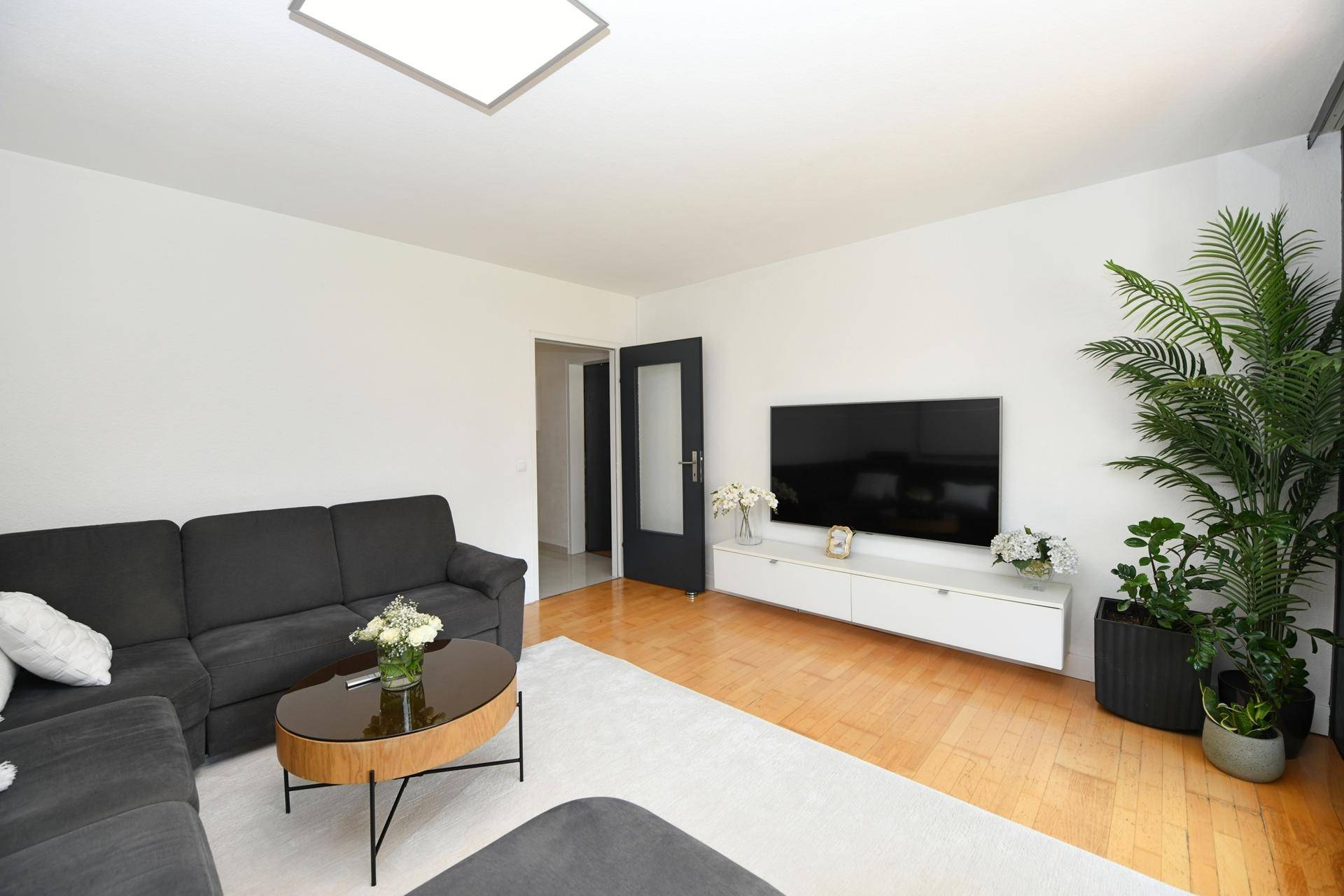Modern eingerichtetes Wohnzimmer mit großem Fernseher, weißem Sideboard und dekorativen Pflanzen.