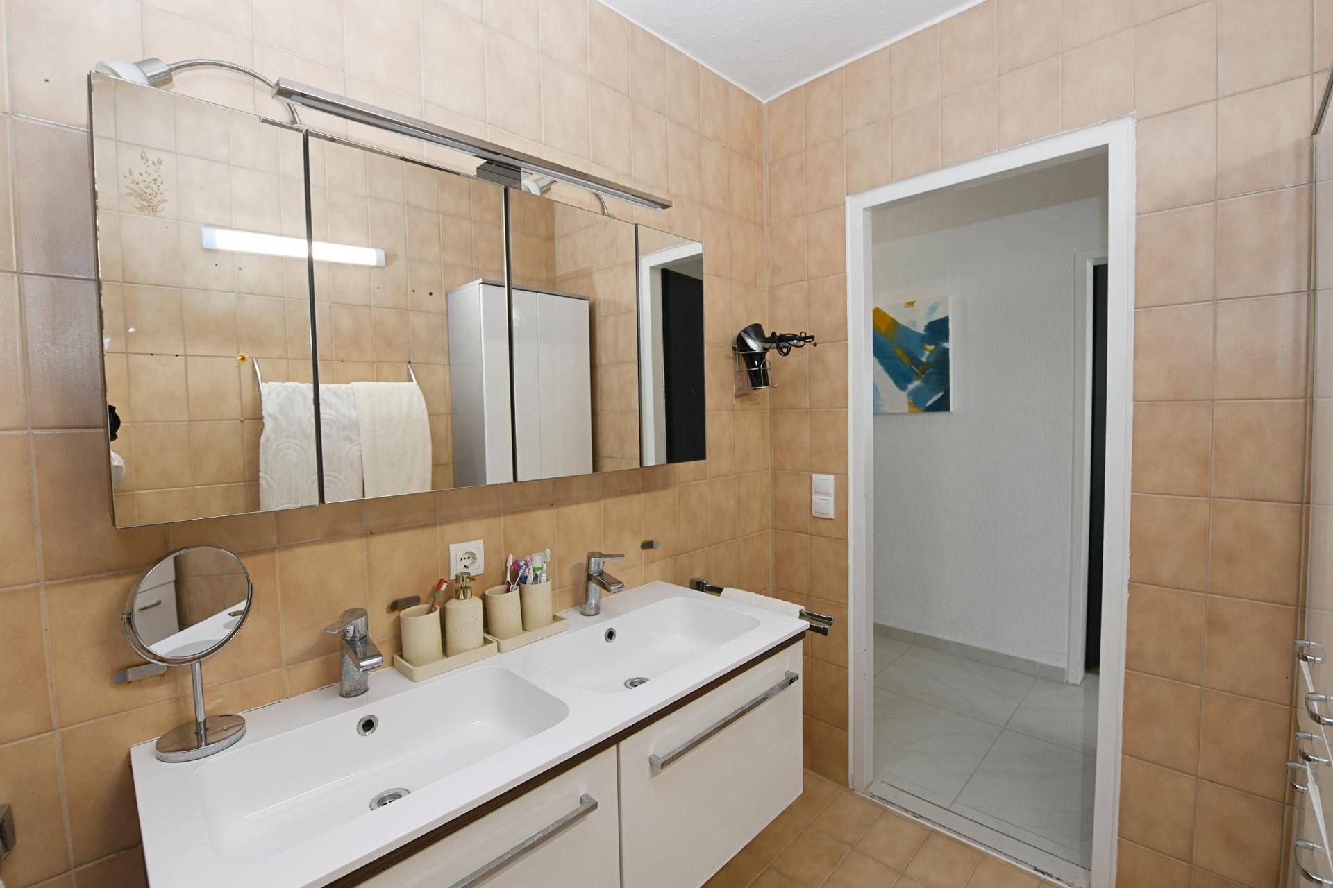 Badezimmer mit Doppelwaschbecken, großem Spiegelschrank und beigefarbenen Fliesen.