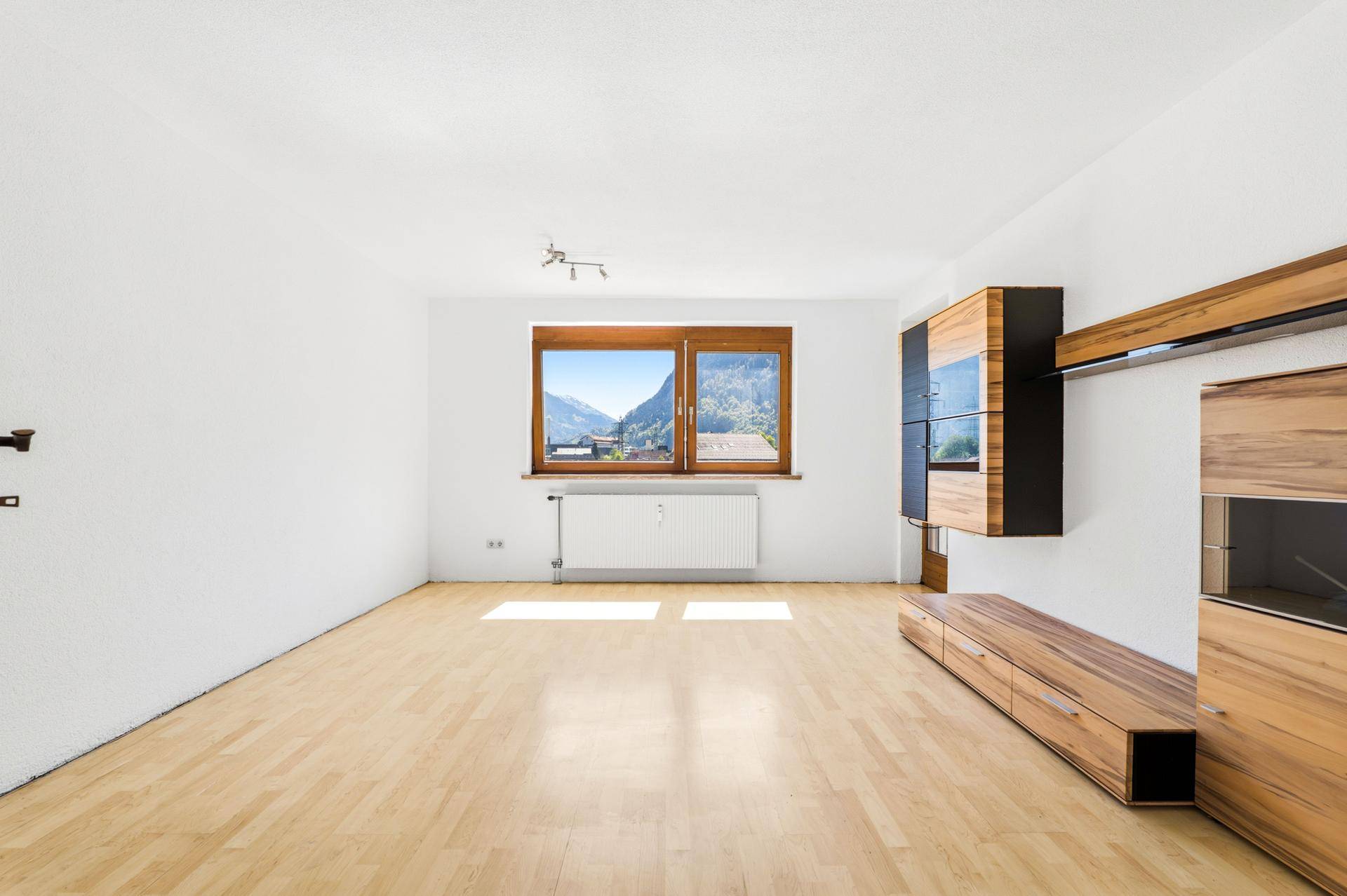 Helles, leeres Wohnzimmer mit Holzboden, Heizkörper und Blick auf die umliegende Berglandschaft.