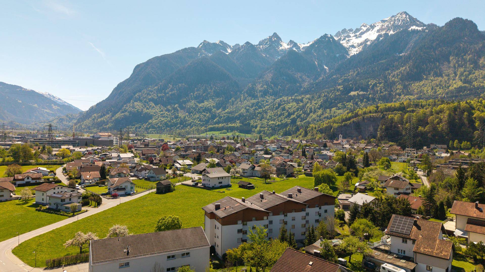 Luftaufnahme einer Wohnsiedlung vor einer beeindruckenden Bergkulisse mit schneebedeckten Gipfeln.