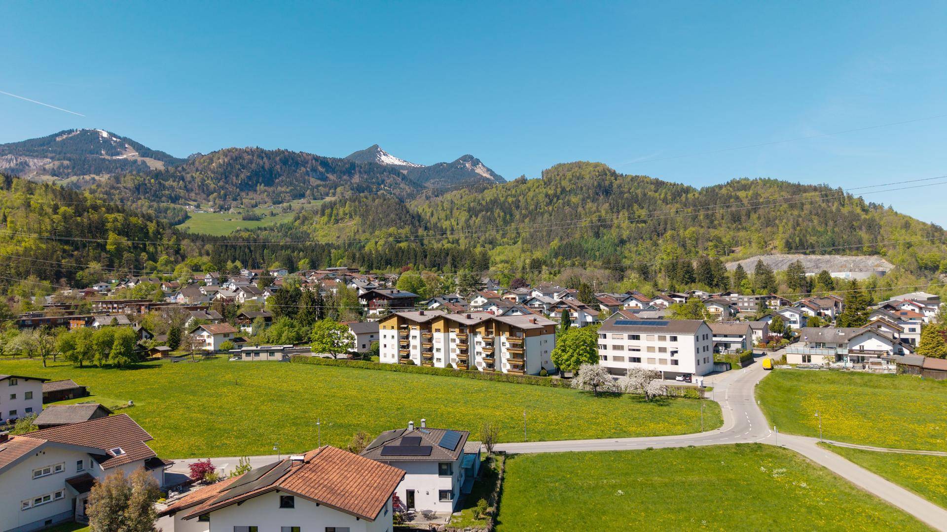 Weitläufige Luftaufnahme einer ländlichen Wohngegend mit Bergen, Wiesen und verstreuten Häusern.