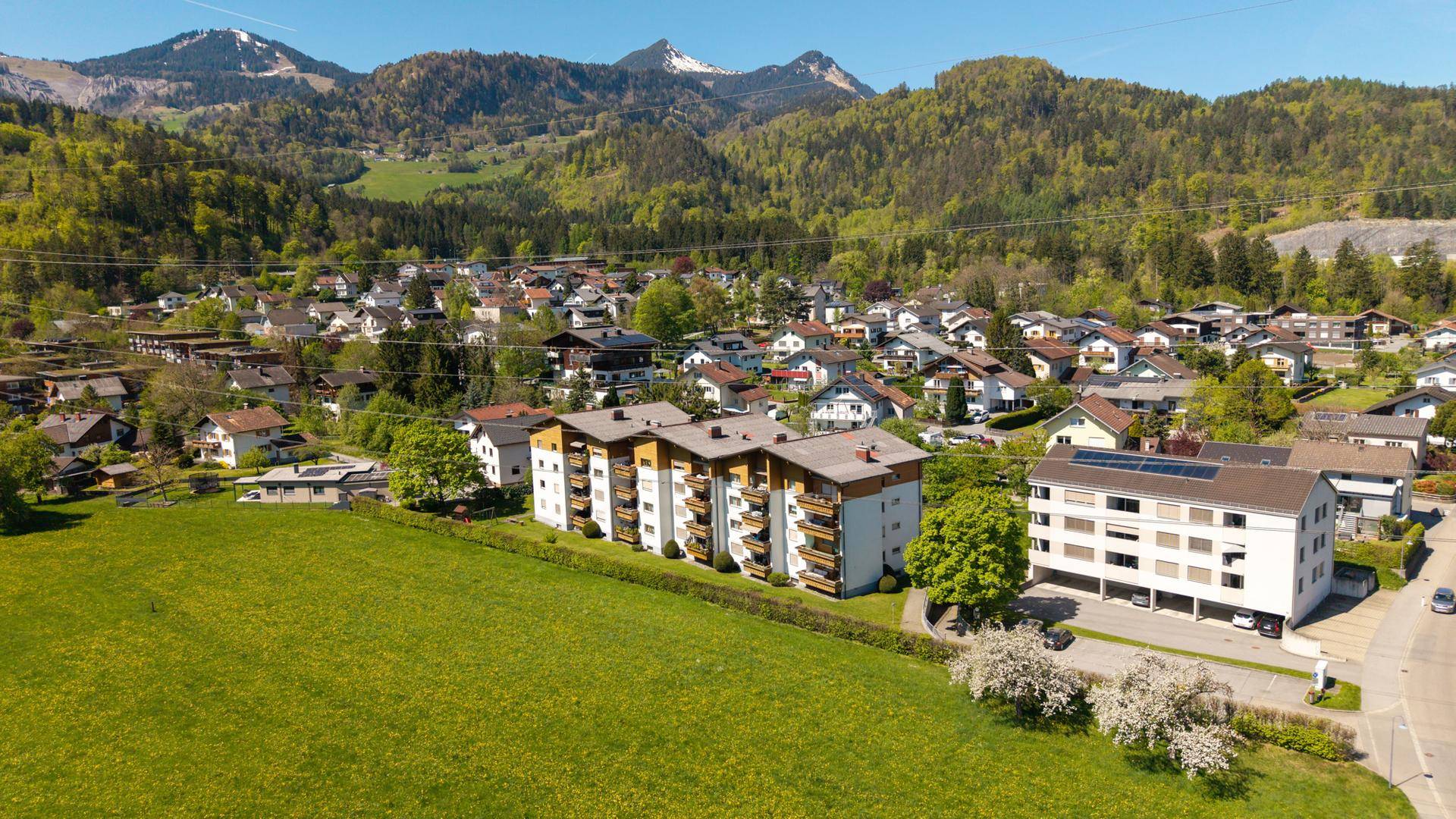 Luftaufnahme einer Wohnsiedlung mit Mehrfamilienhäusern, grünen Feldern und Bergen im Hintergrund.