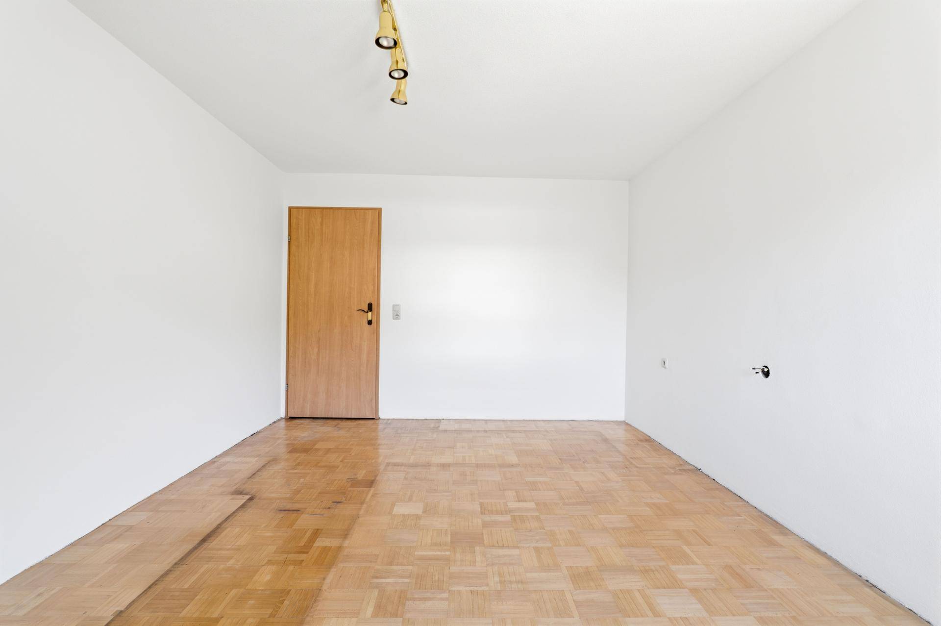 Leeres Schlafzimmer mit Fischgrätparkett, weiß gestrichenen Wänden und Holztür.