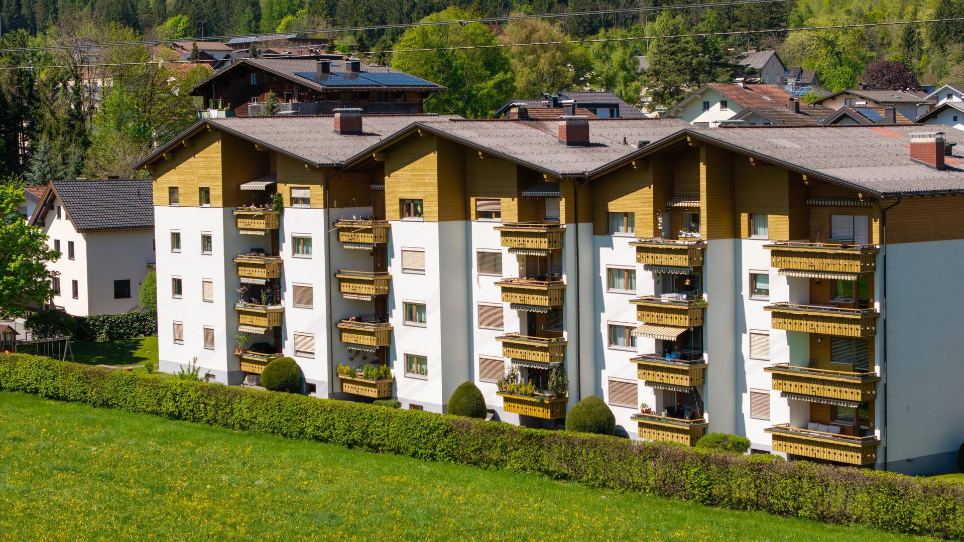 Mehrfamilienhaus mit Balkonen und Holzverkleidung, umgeben von grüner Landschaft und Bergen.
