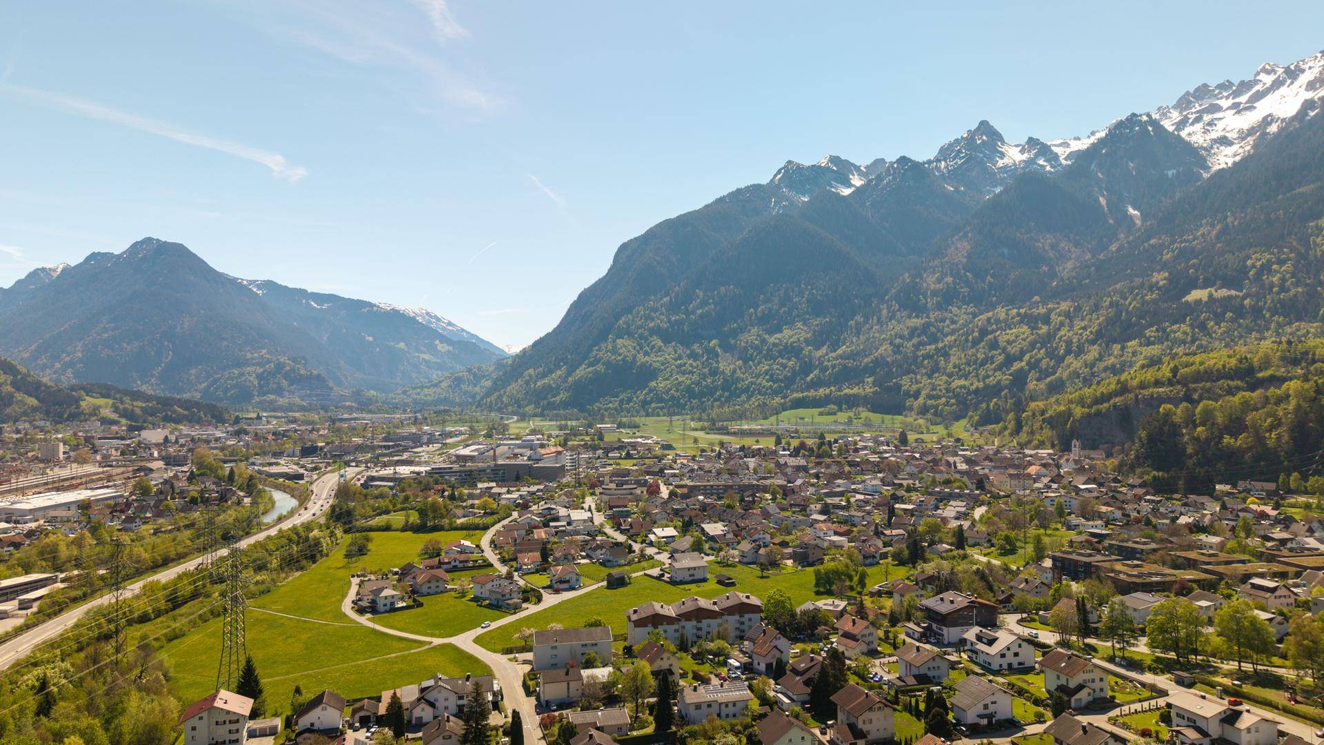 Weitwinkel-Luftaufnahme einer Stadt im Tal, umgeben von grünen Bergen und einem Fluss.
