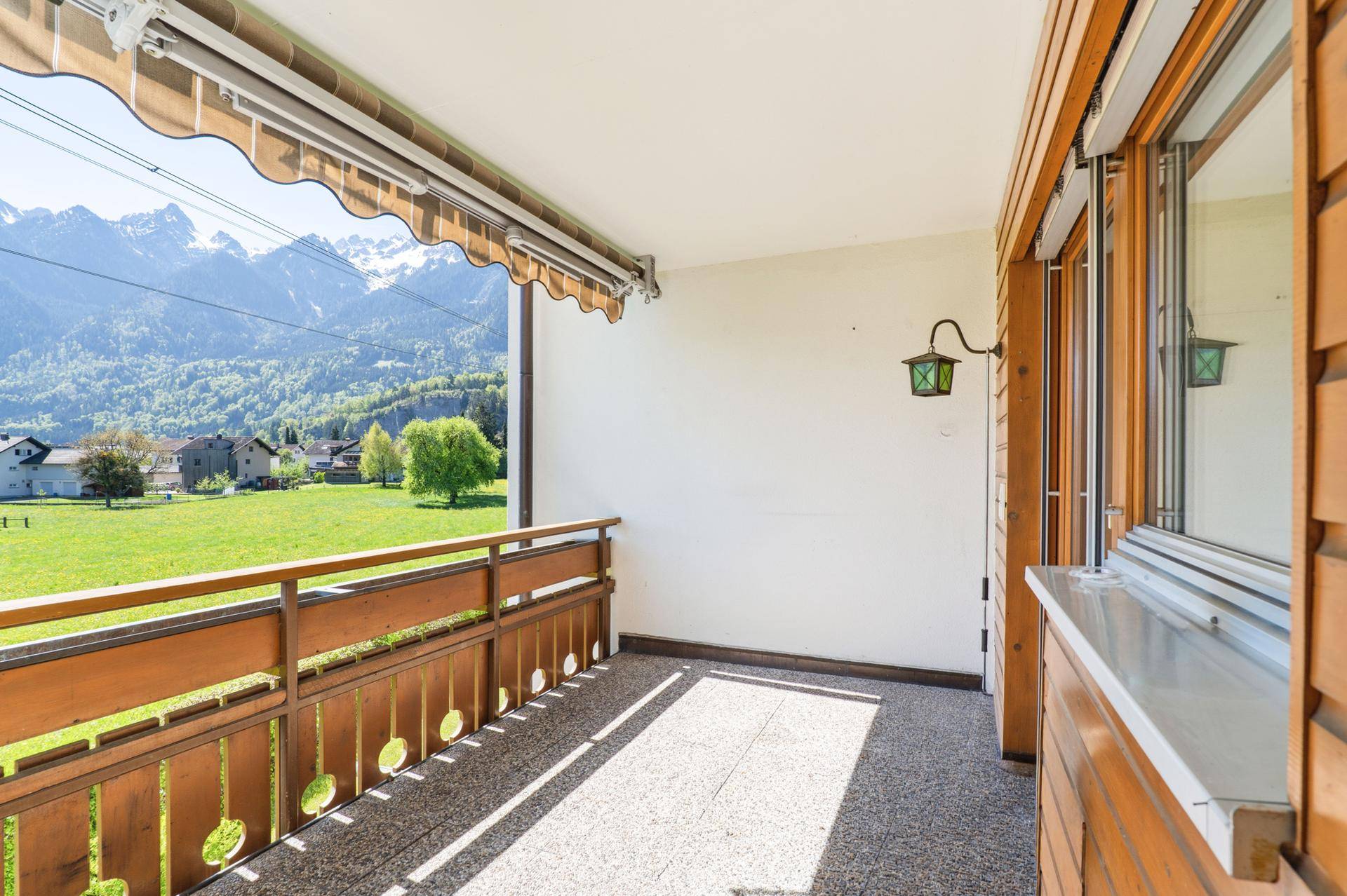 Sonniger Balkon mit Markise, Holzelementen und weitem Blick auf die Berge und grüne Wiesen.