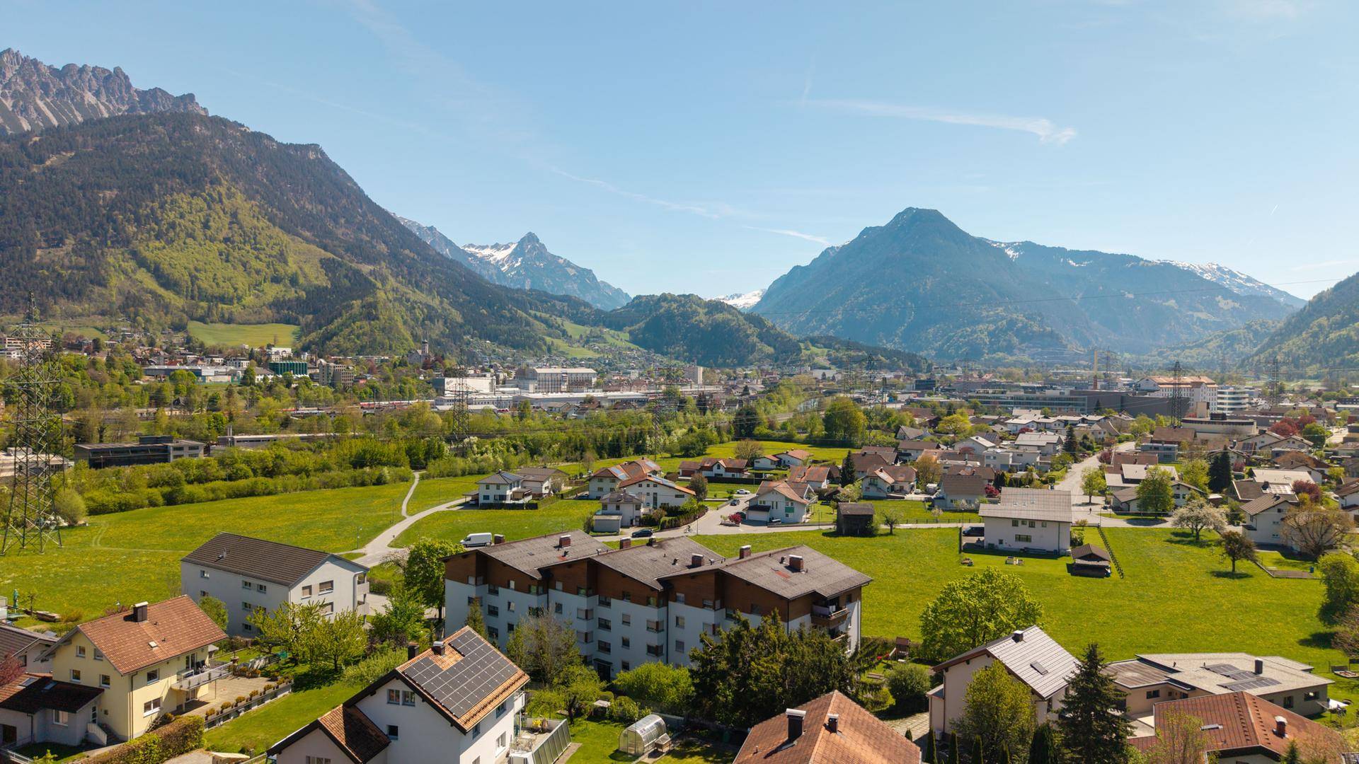 Panoramablick aus der Vogelperspektive auf eine Siedlung mit Bergen, Feldern und Häusern.