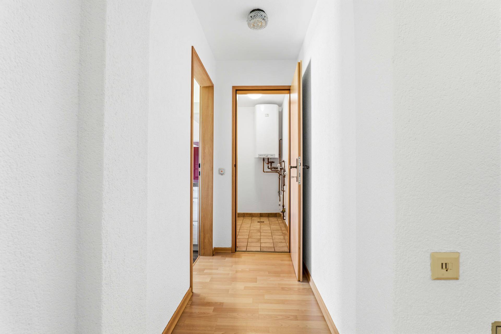 Heller Flur mit Holzboden und Blick auf einen Raum mit Warmwasserboiler und Fliesenboden.